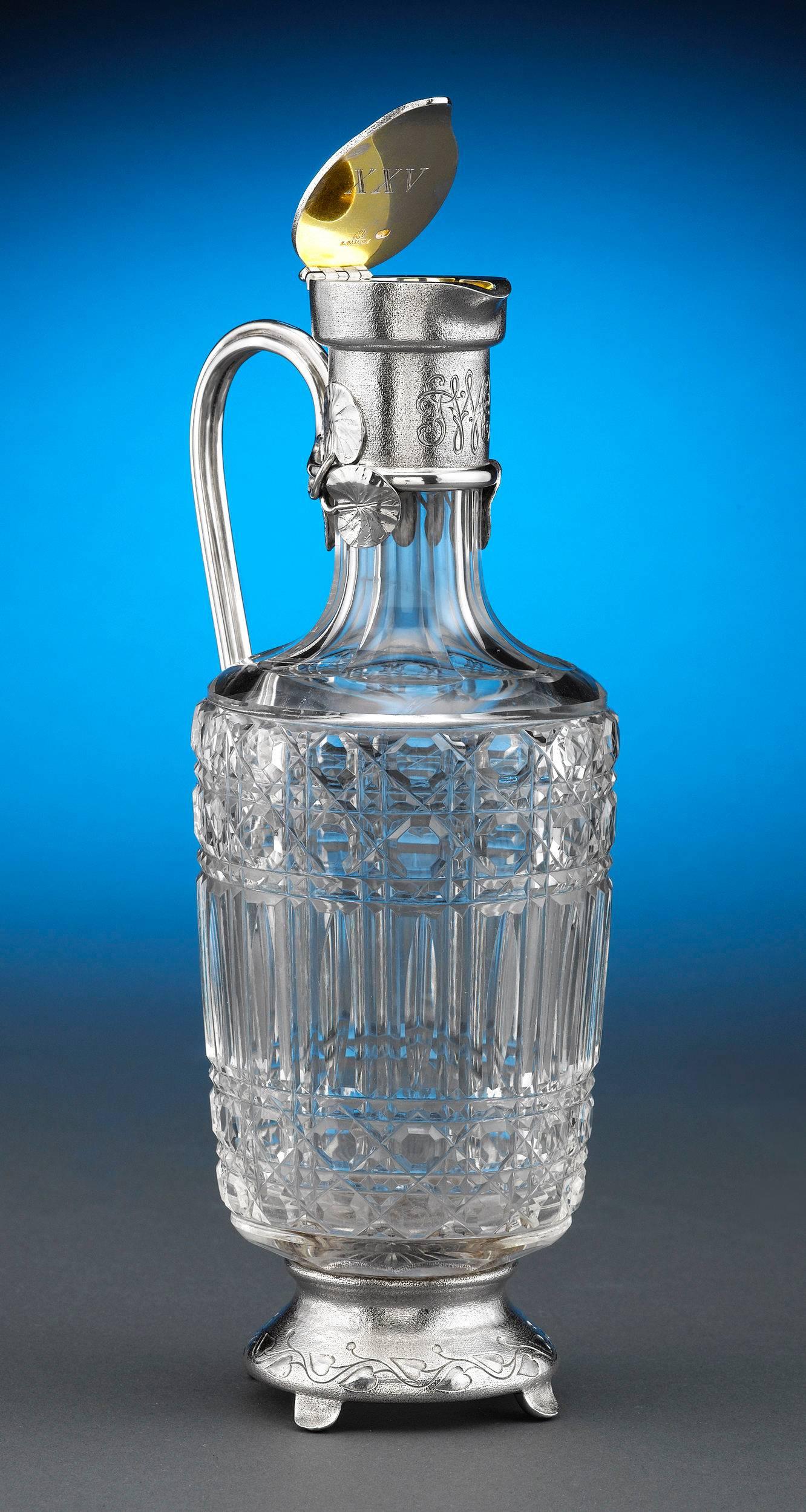 This incredible claret jug by Fabergé is a study in balanced design. Crowned by a silver lid adorned with a lily pad motif, the body is crafted of intricate cut-glass and bears a wonderful cane pattern, while its silver footed base is chased with a