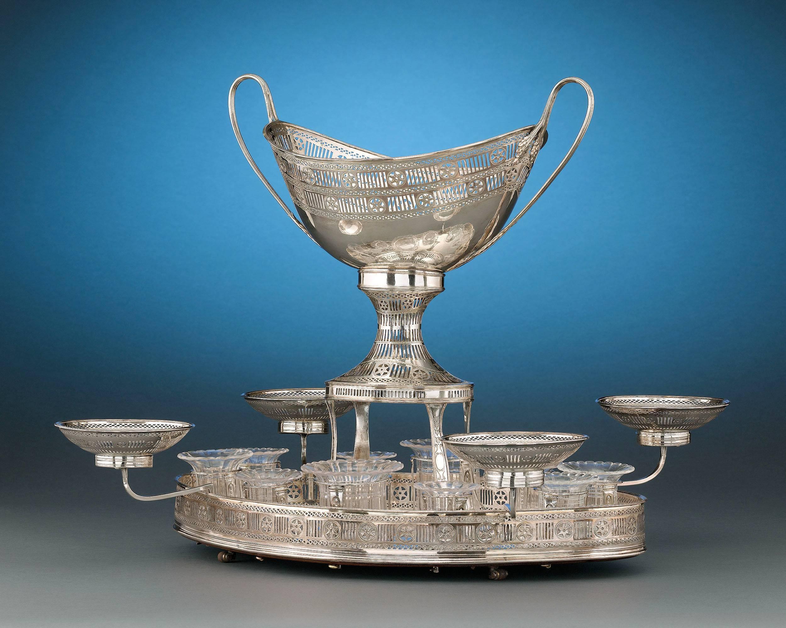 The perfect complement to a fine dining affair, this George III-period rolling dessert stand is crafted of desirable Sheffield silver plate. In this modified version of an epergne, this centerpiece features an urn-form basket encircled by pierced