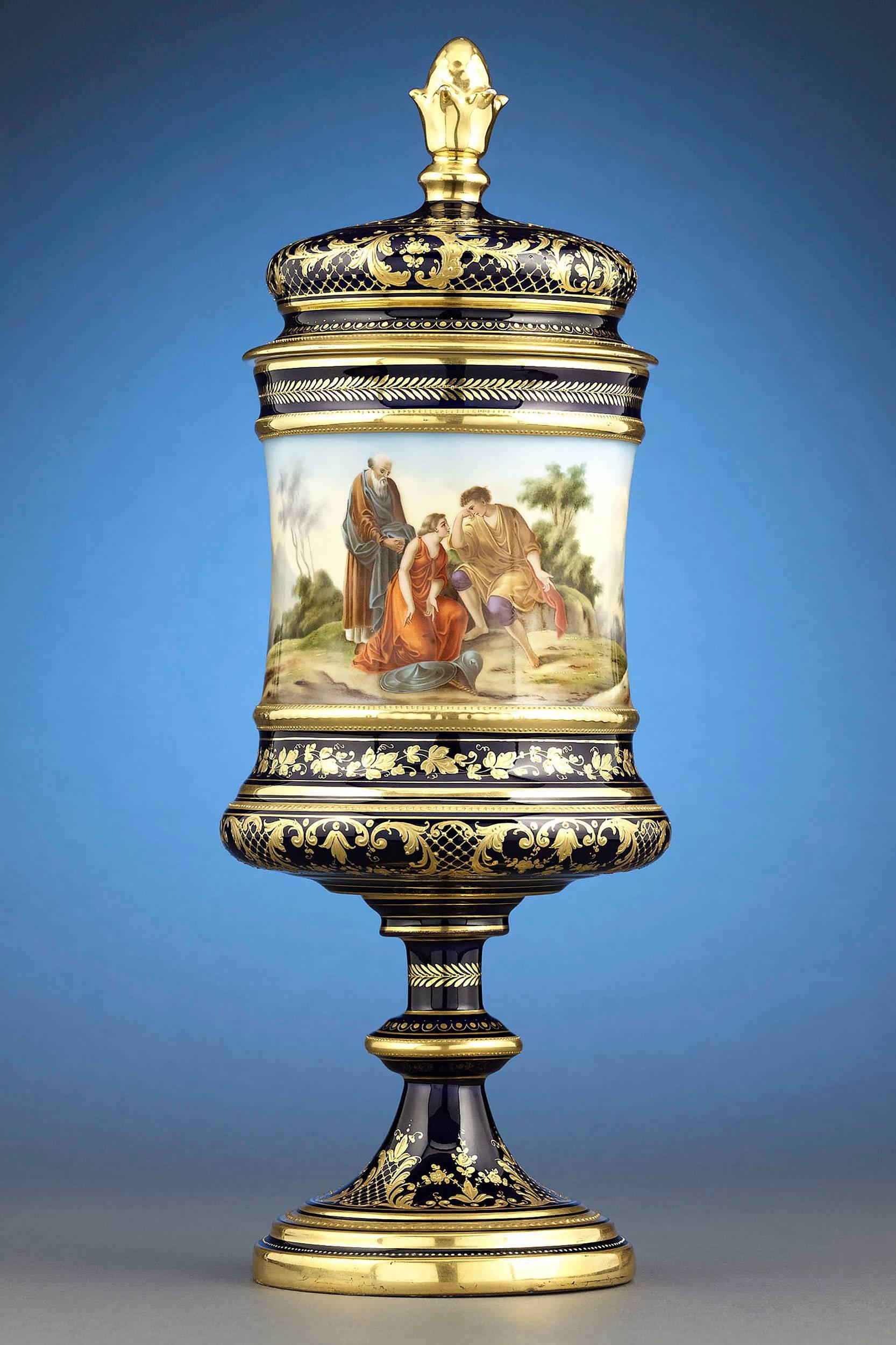 A beautiful Royal Vienna Porcelain urn showcasing the firms renowned rich, cobalt blue background contrasted by lush gilt accents. A classical figural scene wraps around the entire vessel, depicting Cupid being restrained from releasing another