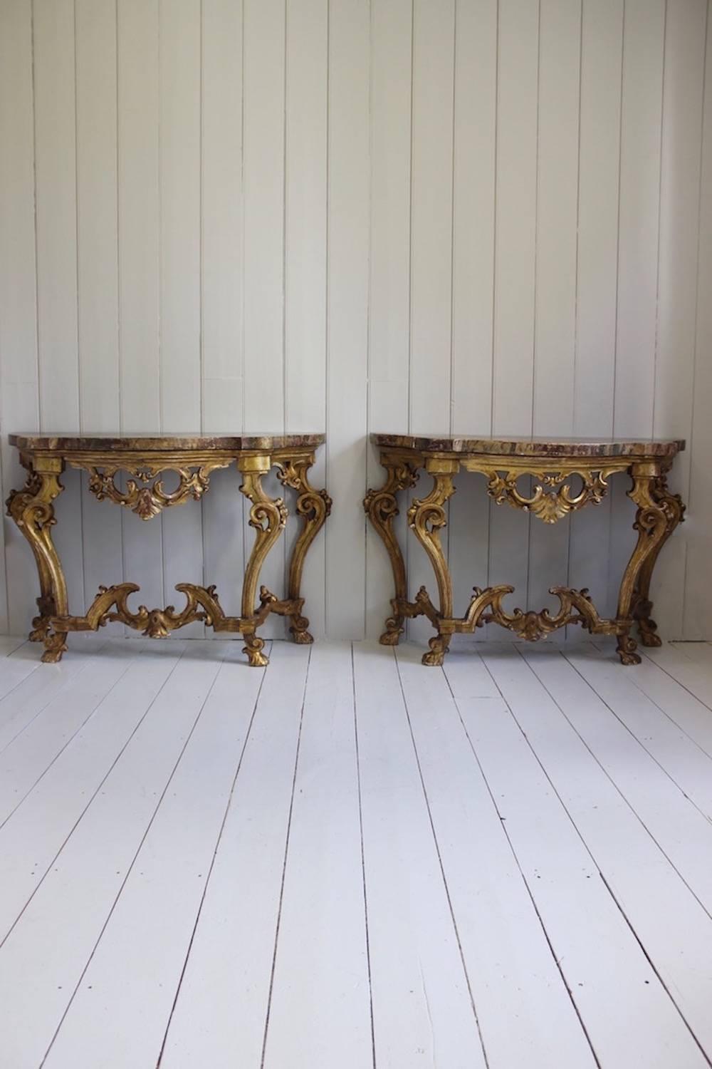 A great pair of carved giltwood console tables with faux marble tops, of serpentine form with exuberantly shaped bases, in the Rococo style. The oil-gilding is richly clouded and burnished and the painted tops offer a subtle contrast, Italian, 19th