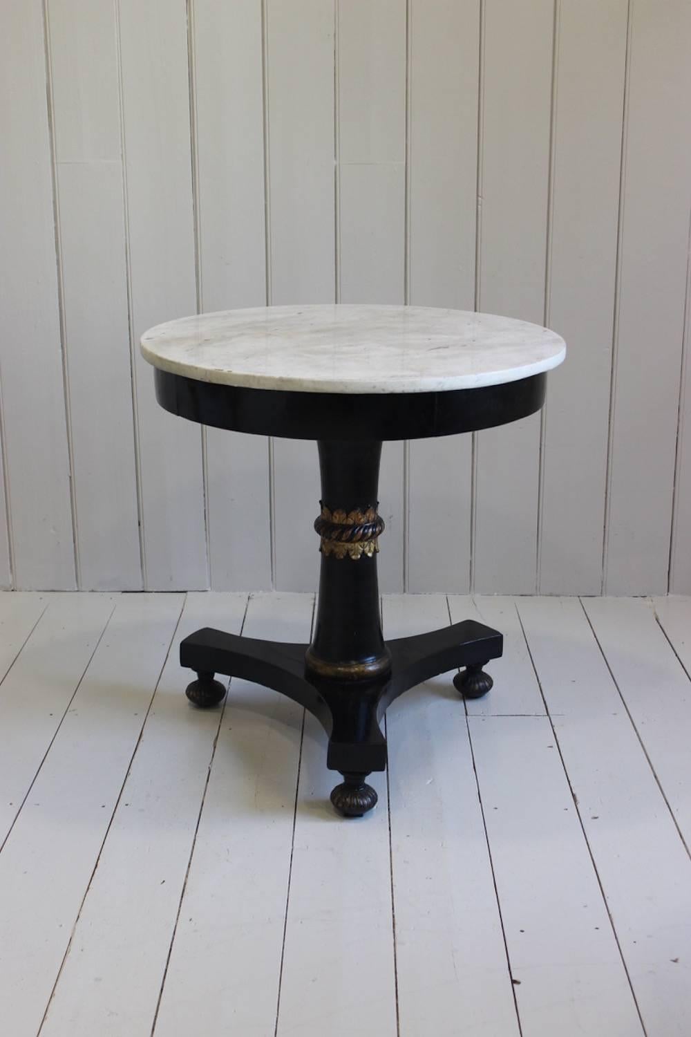 A very smart and stylish ebonized and parcel-gilt gueridon or small centre table with its original white marble top and triform base with bun feet, the column with a spiral-bound decorative stiff-leaf motif, Italian, early 19th century.