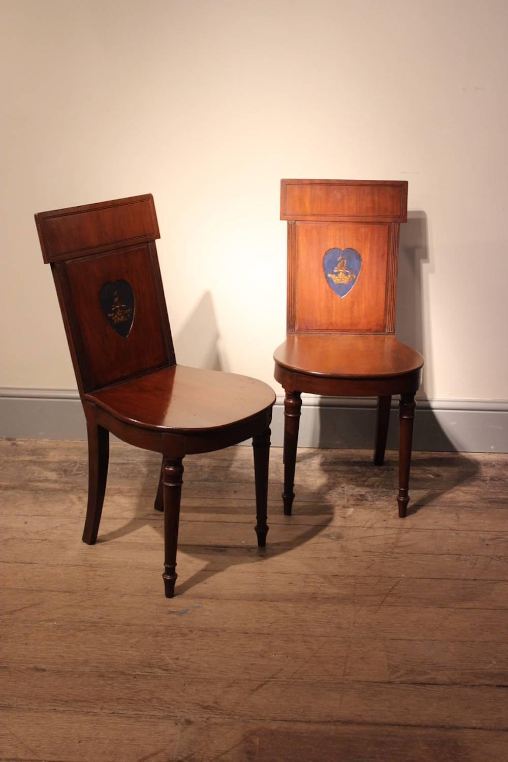 A smart pair of Regency period mahogany hall chairs with solid tablet backs, centred by painted armorial heart-shaped cartouches, with rounded seats and turned tapering legs, English, early 19th century.