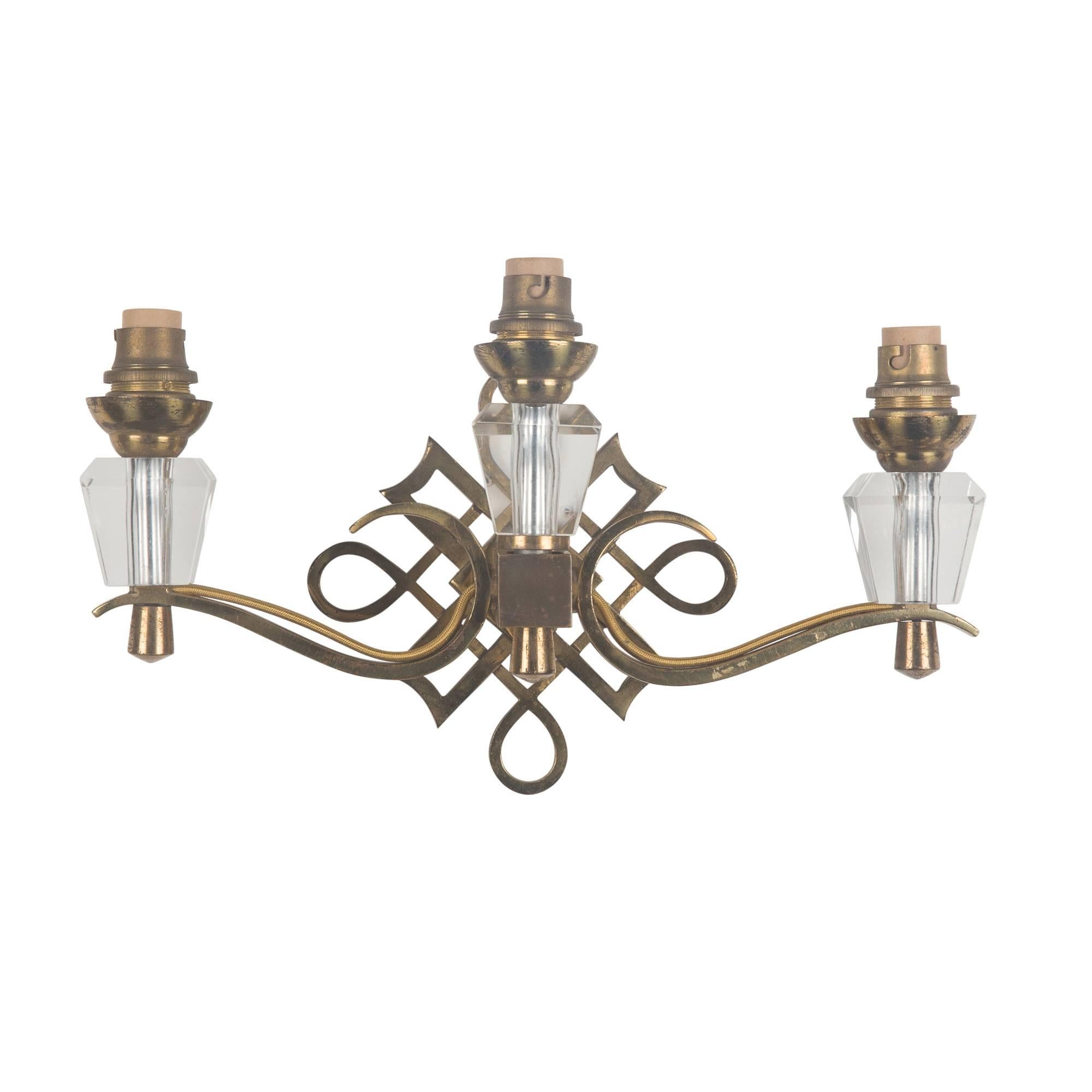 Pair of two arm, three light wall sconces, in bronze, with swirled decorative back element, faceted crystal socket undermounts, in the style of Jules Leleu, France, 1930s.Measures: Height to top of socket 6 in, width 12 1/2 in, depth 4 in.