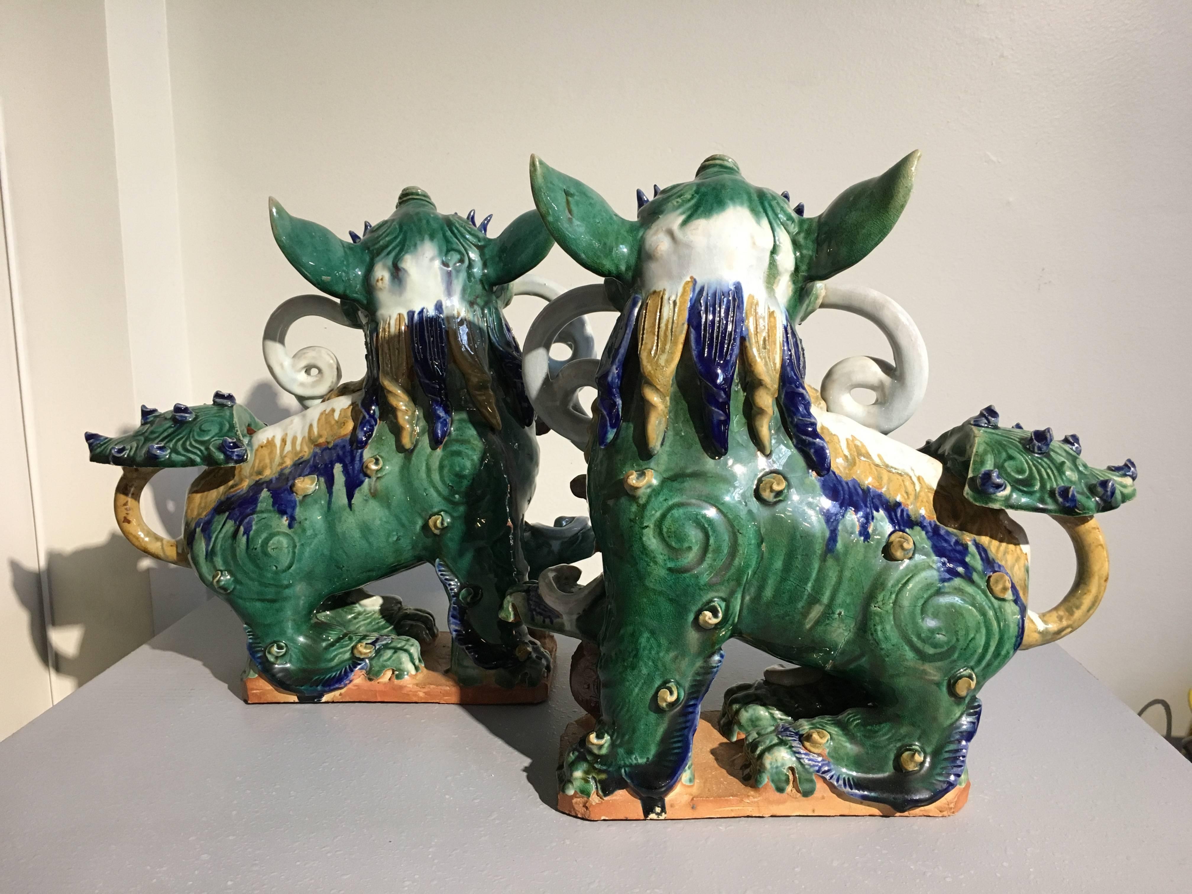 A delightful pair of Chinese pottery foo dogs or lions glazed in vibrant green, with blue, yellow and white highlights. 
Both are portrayed standing, with heads turned and holding streaming banners adorned with flowers in their mouths. They have