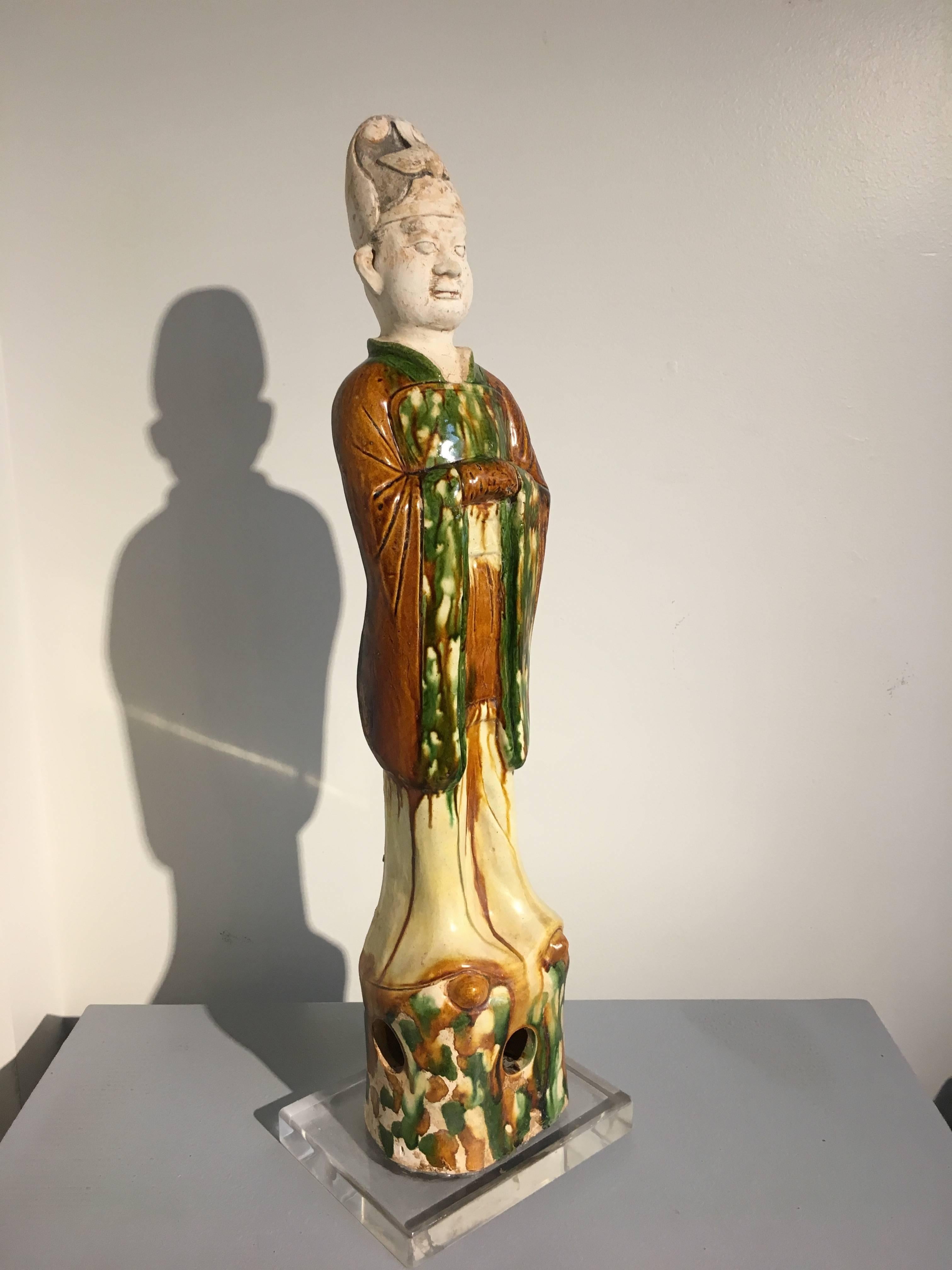 A Tang dynasty sancai (three color) glazed pottery model of a Taoist official, TL tested by Oxford Authentication. The figure is portrayed standing upon an openwork base in an attentive position, dressed in loose fitting robes, his clasped hands in