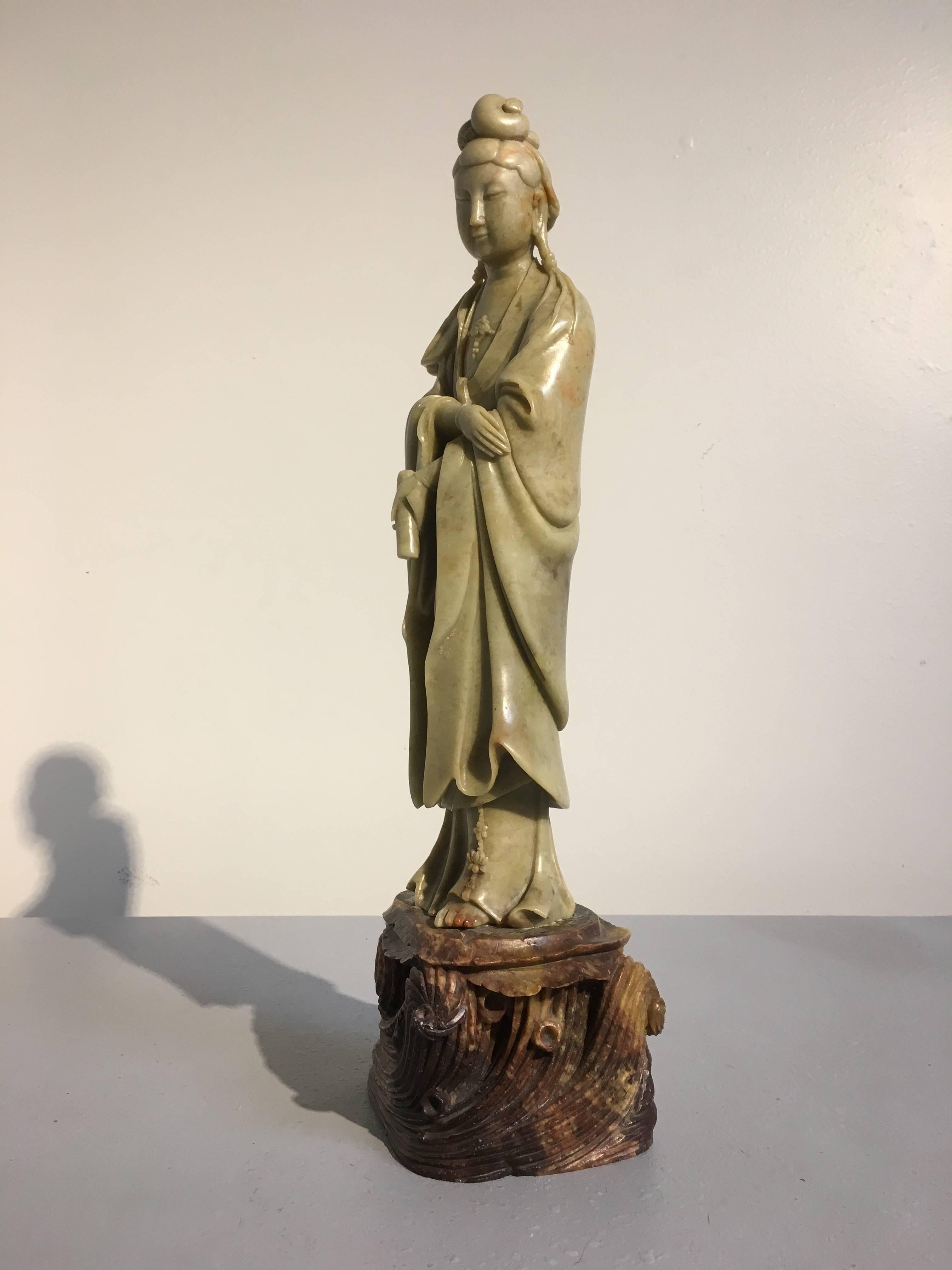 A sublime Republic Period carved soapstone figure of the Chinese Goddess of Mercy, Guanyin. The Buddhist Bodhisattva of Compassion stands upon a separately carved rocky outcrop surrounded by crashing waves. Her head slightly bowed, a demure smile