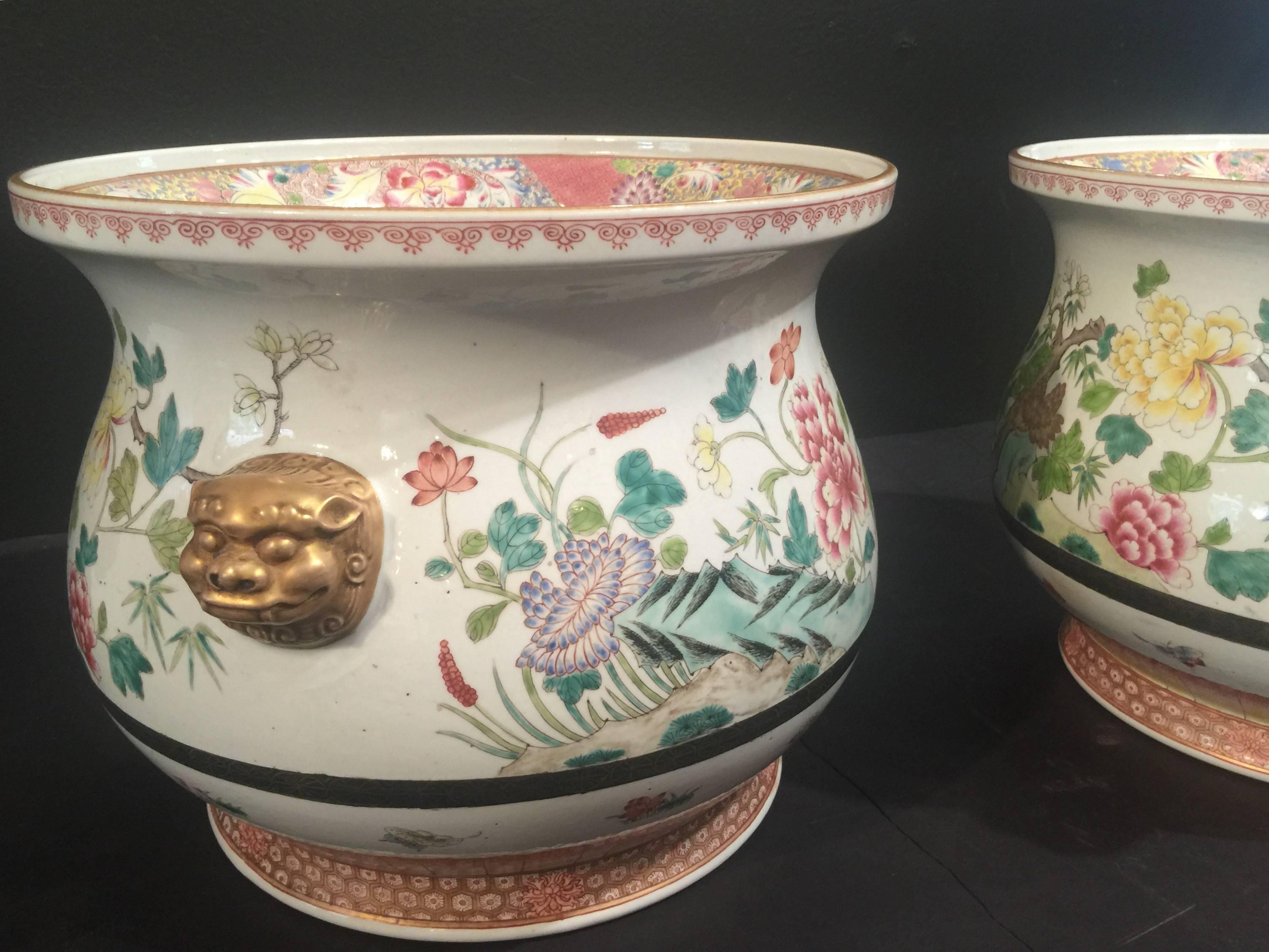 A stunning pair of Chinese porcelain cachepots, finely painted in famille rose enamels. The bulbous body features a Traditional Design of blossoming flowers, including peony, lotus, chrysanthemum and dogwood, with a pair gilt lion mask handles. The