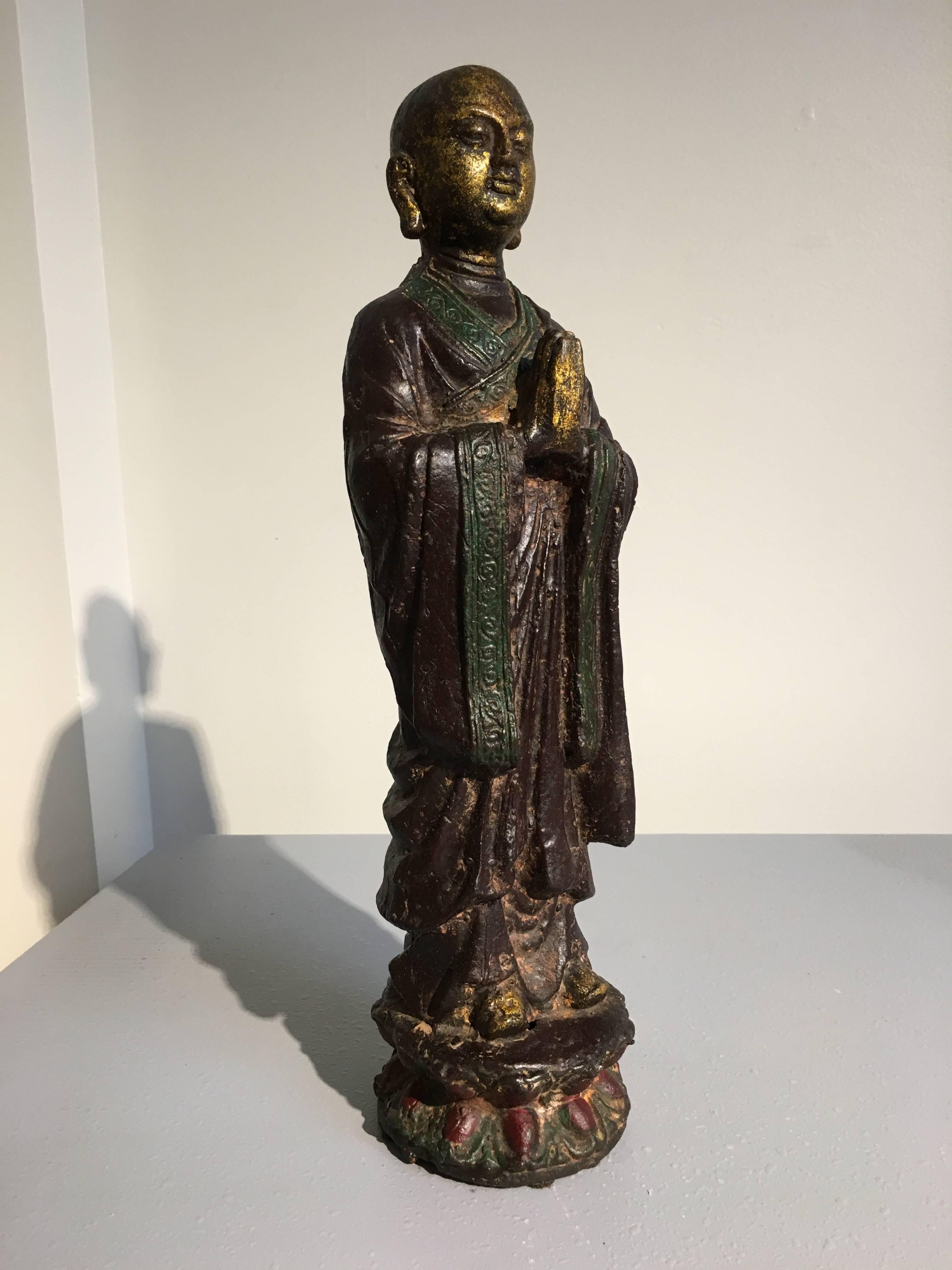Ananda is portrayed in a typical fashion, as a youthful monk with a shaved head and dressed in simple robes. He stands upon a double lotus pedestal with hands clasped in front of him, eyes cast downwards, a serene expression upon his face. 
The