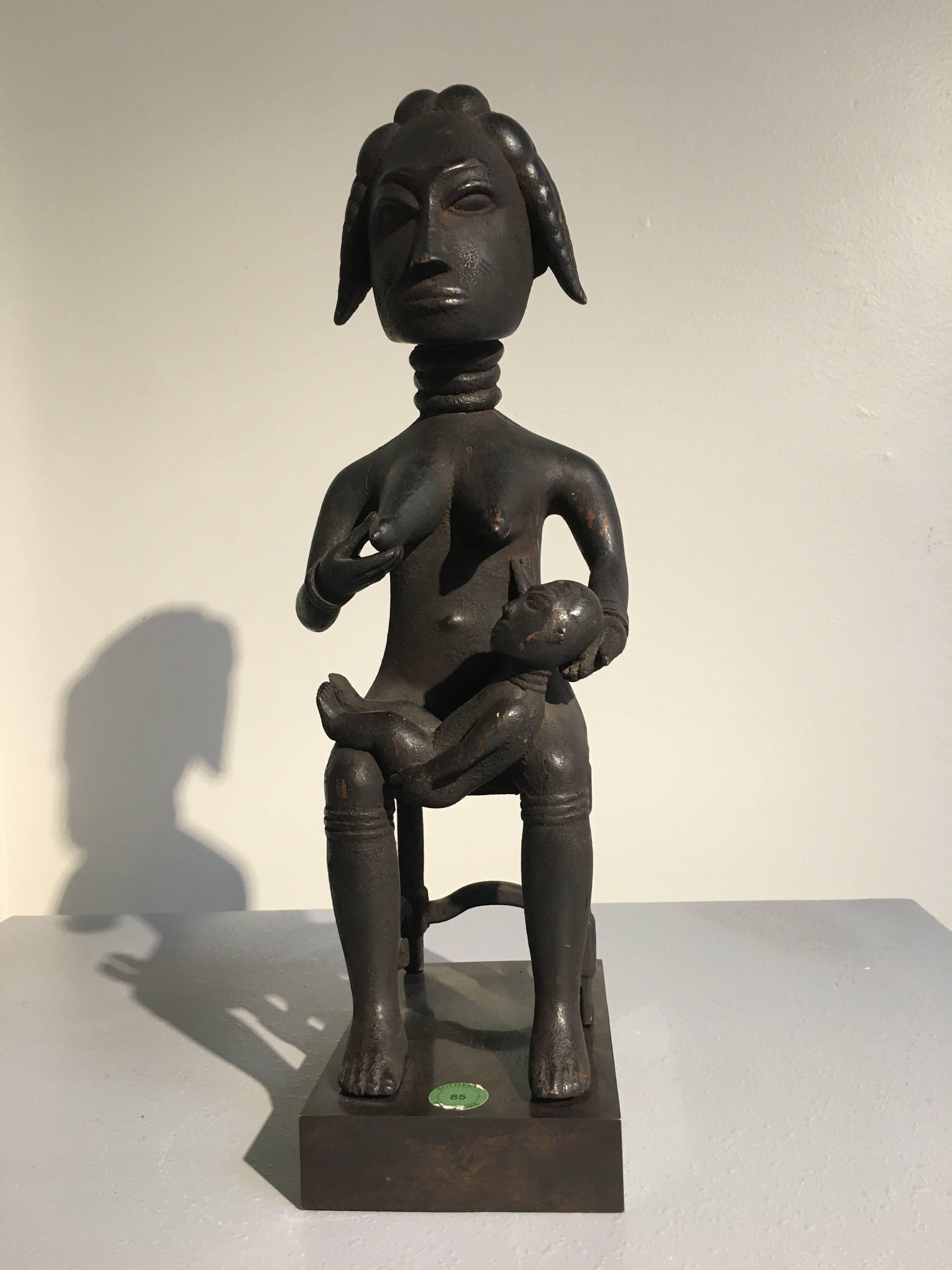 A fine Ashanti maternity figure by the master carver Osei Bonsu. The mother figure is seated on a simple backless chair. An infant lays calmly in her lap, one arm reaching towards a pronounced breast. She cradles the child gently with one hand, the