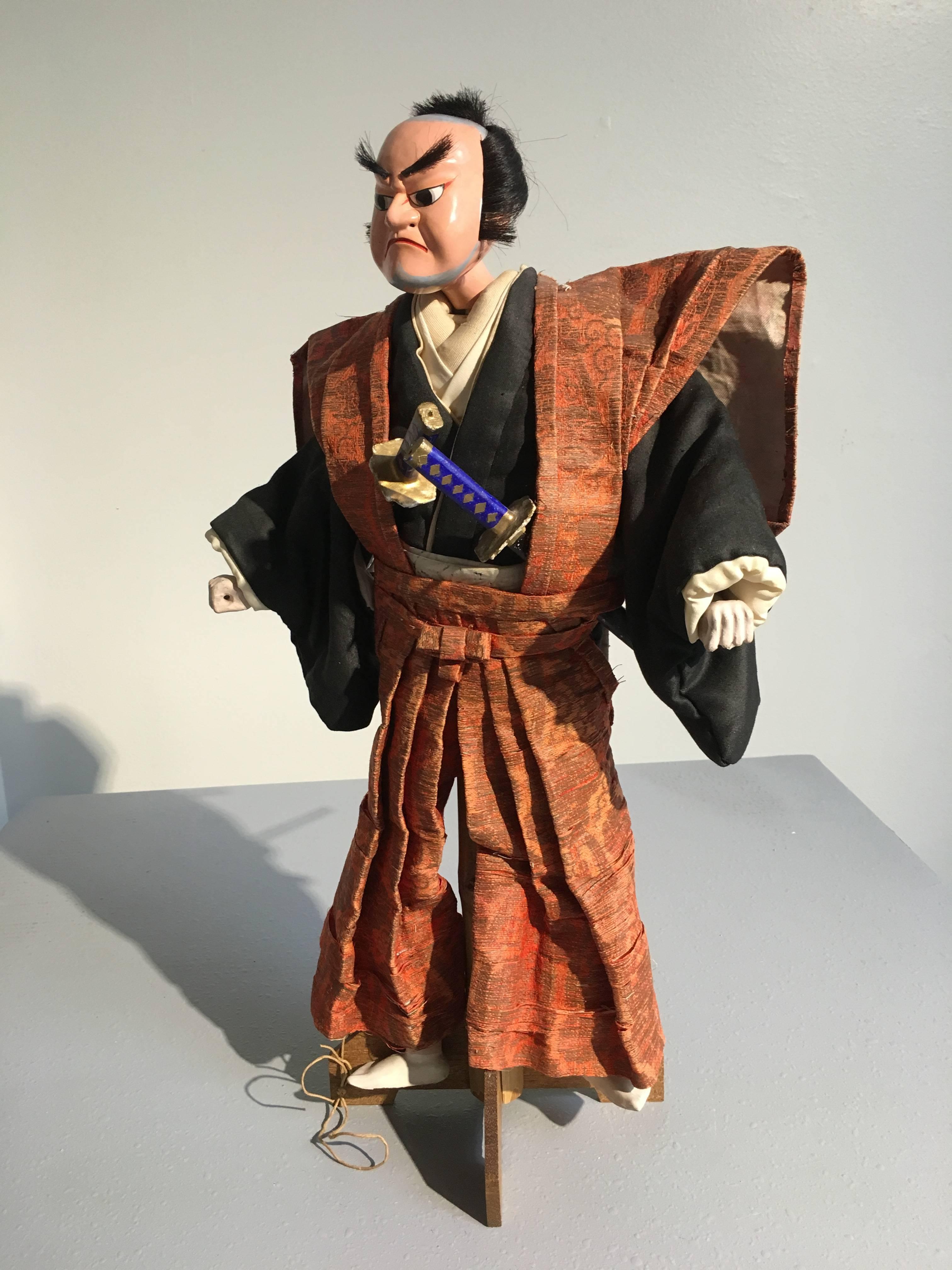 Created for Bunraku theatre, this Bunraku, known in Japan as a Ningyo Jorurui, depicts a samurai warrior. He is dressed in full kamishimo, with a kataginu and hakama of rich brocade featuring gold thread on a coral ground. A pair of removable swords