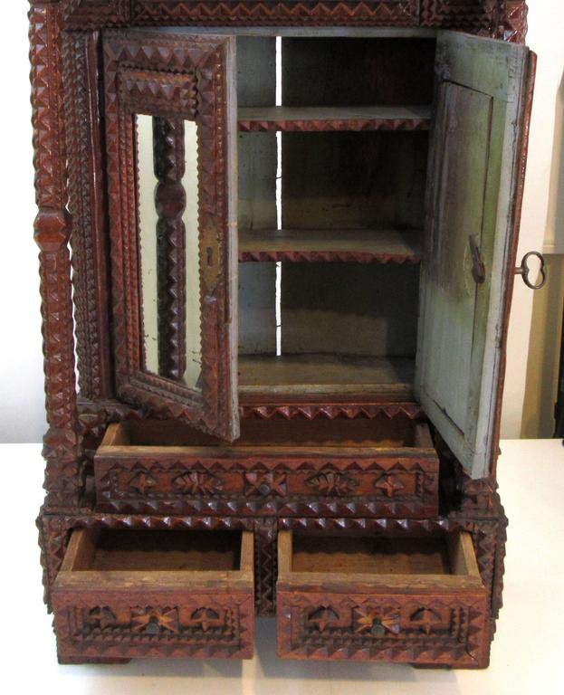 An antique Tramp Art hanging cabinet with mirrored panel doors that open to a single shelving unit, over one long drawer over two short drawers.
