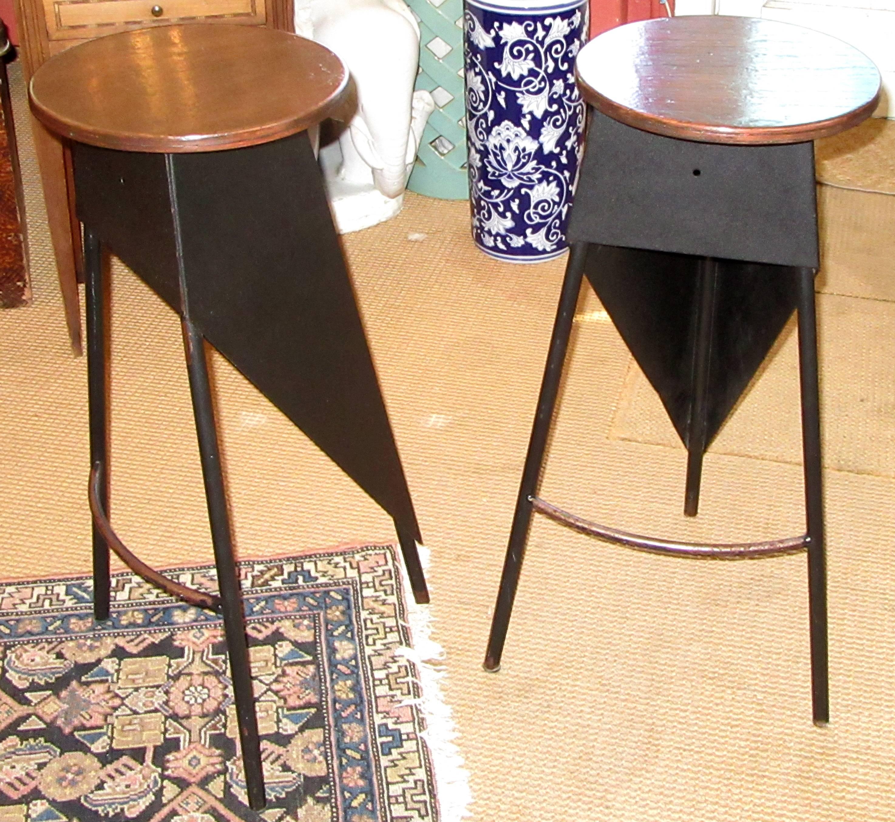 A pair of black painted steel stools with round wood seats. Geometric design with a rounded footrest, topped with stained and lacquered pressed wood seats.