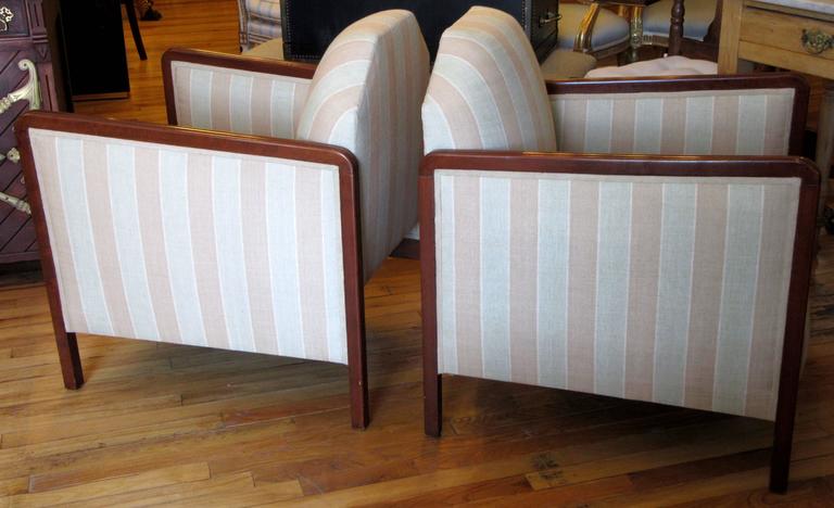 A pair of slender, deep seated Art Deco era club chairs. Geometric form back and continuous leg to leg wood trimmed arms.