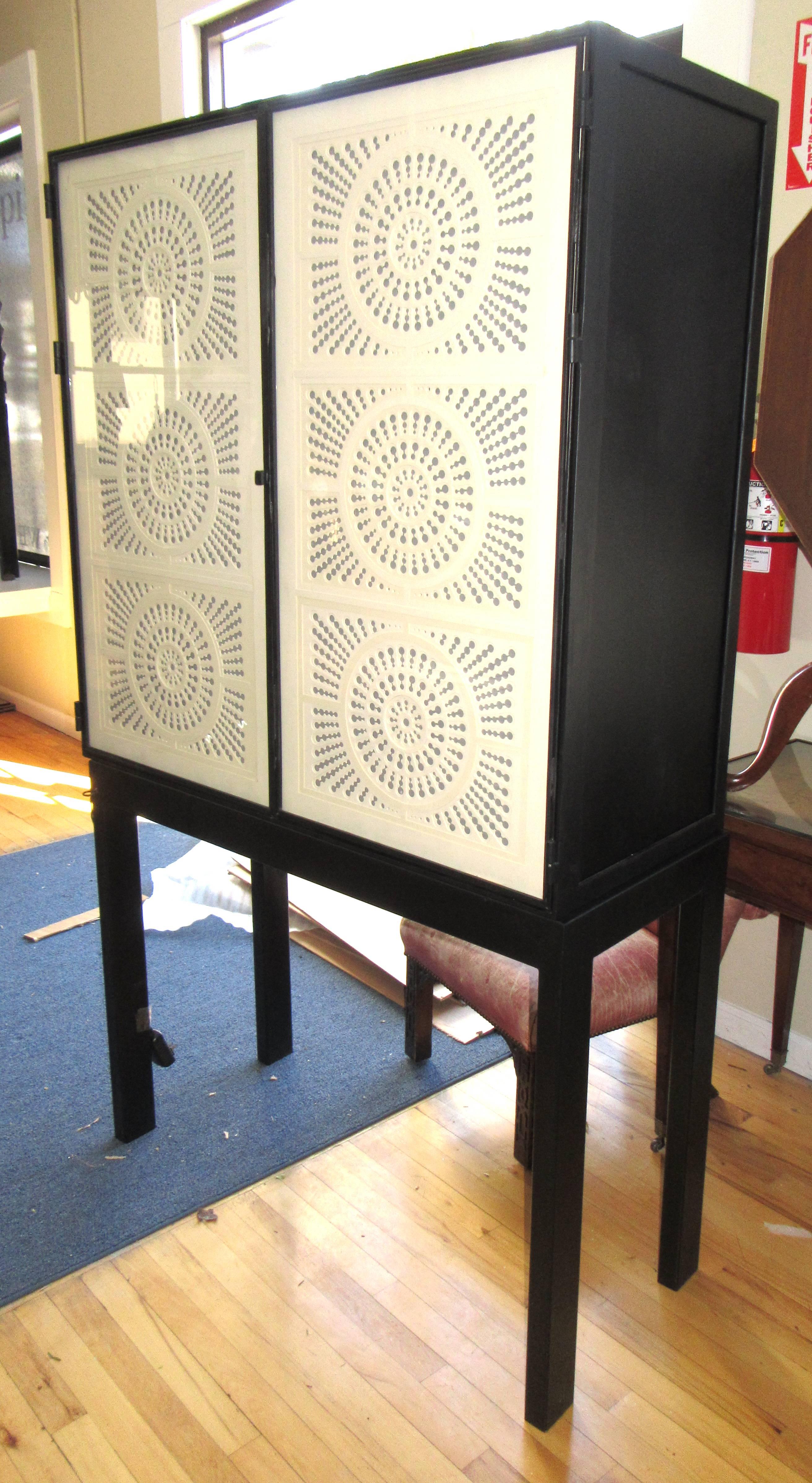 An iron frame cabinet on stand. The doors are glass, lined with intricately cut-out paper. The cut-out design allows the fabric lined interior to glow through. The shelves are removable glass. The cabinet is wired to light from the interior.