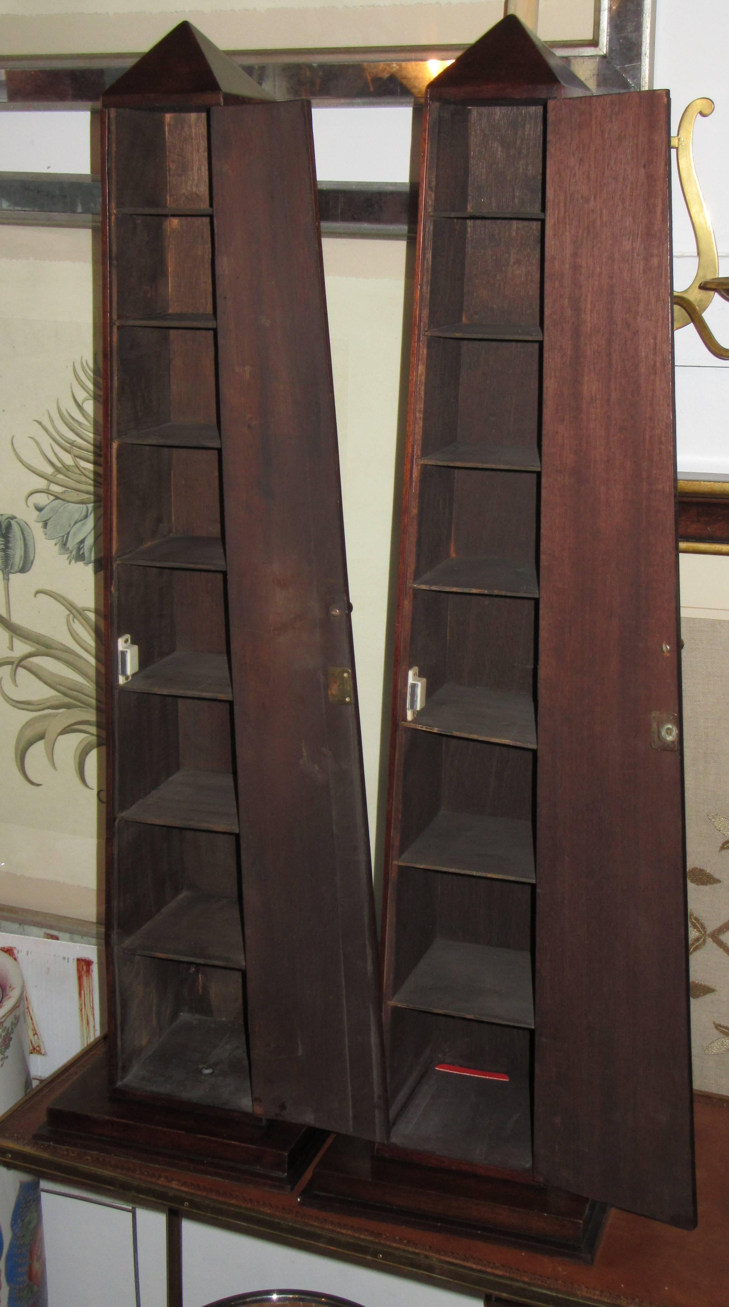 A pair of mahogany obelisks on platforms. One side opens to reveal graduated shelves.