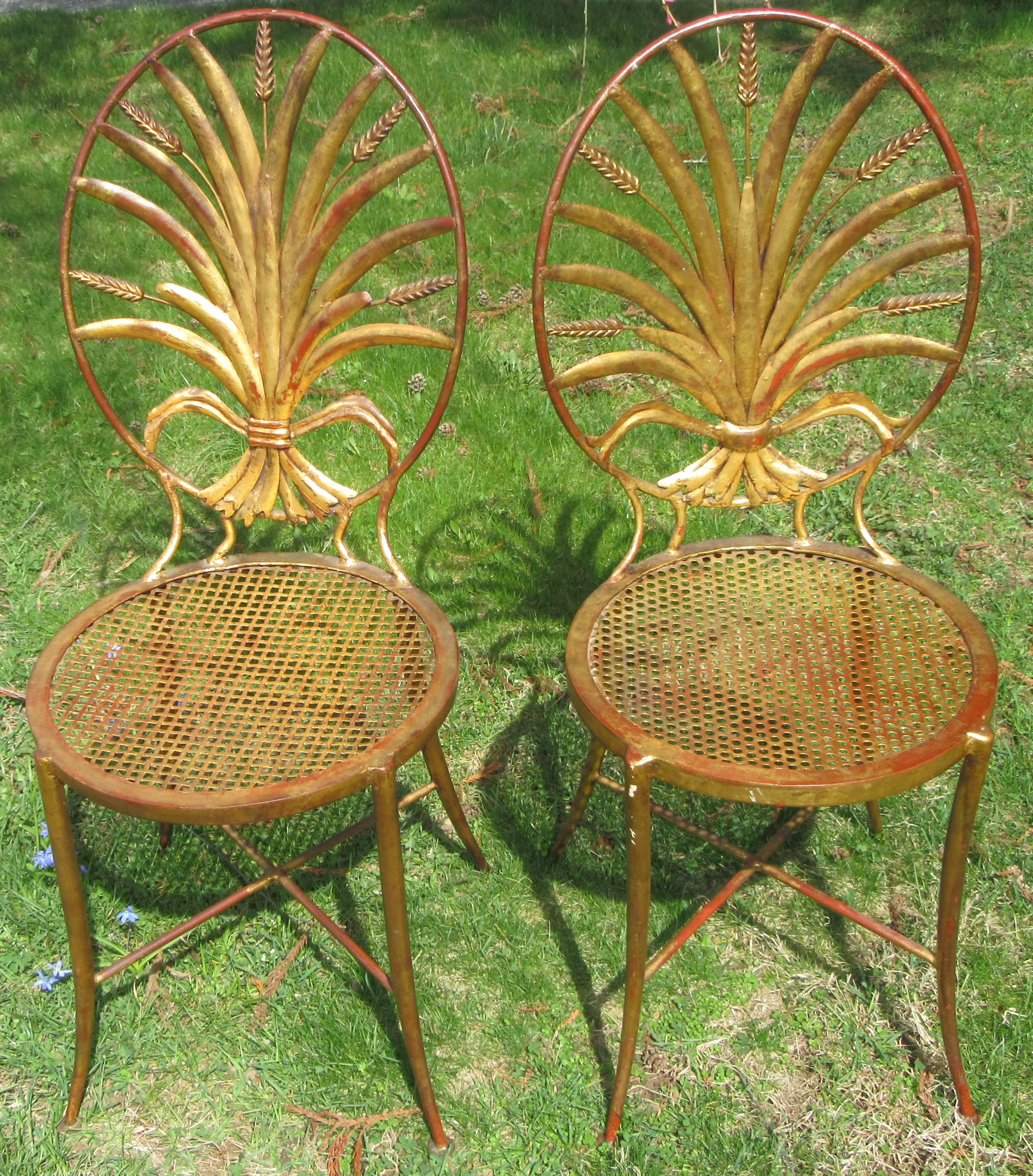 A pair of metal bistro chairs with a gilded finish. The back is an oval containing a fanned out sheaf of wheat design.