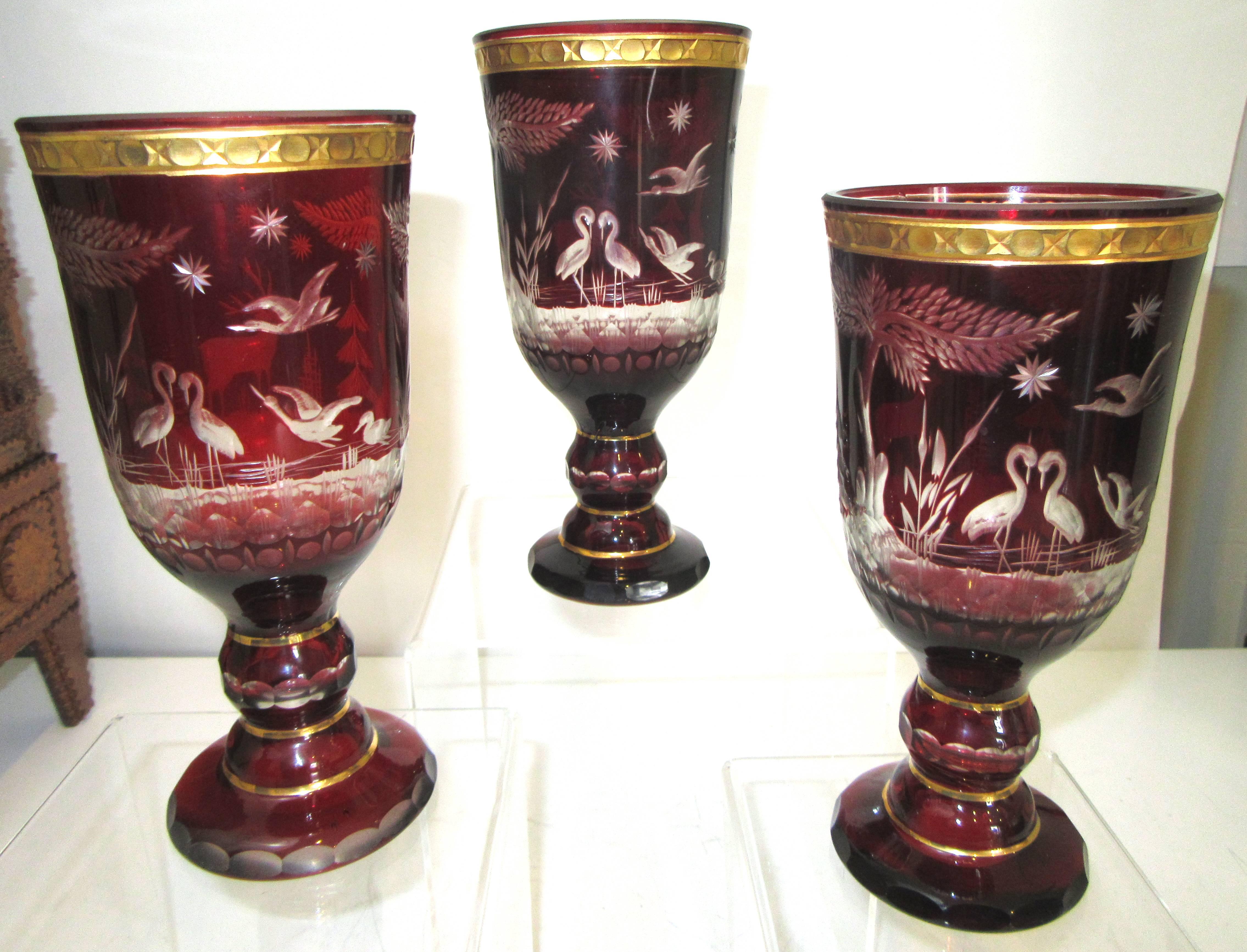 Three tall and substantial ruby red goblets that are cut to clear depictions of nature and wildlife. Each has a gilded rim.