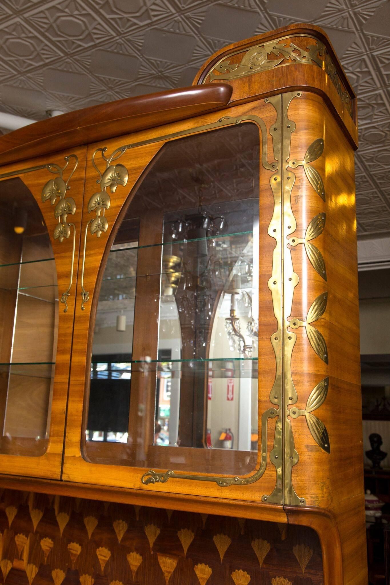 A two-part early 20th century French cabinet. The top of mixed fruitwoods with marquetry, mirrored panels and glass doors above a marble base alcove. The lower section with two drawers opened by solid brass handles, over a two-door cupboard. The