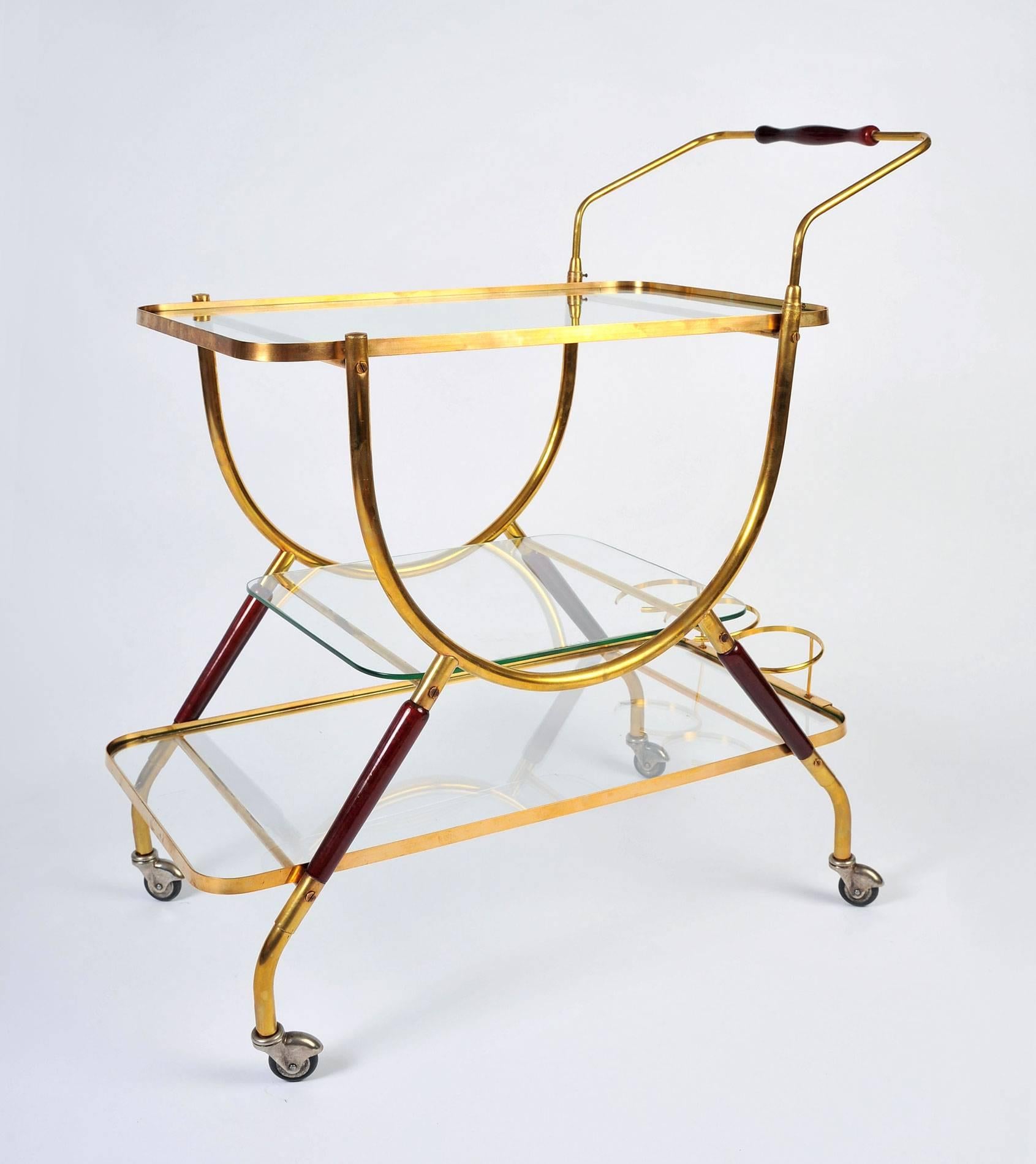 Elegantly practical French bar cart with brass frame, deep red wooden leg detail and matching red handle. Three glass shelves and three bottle holders.