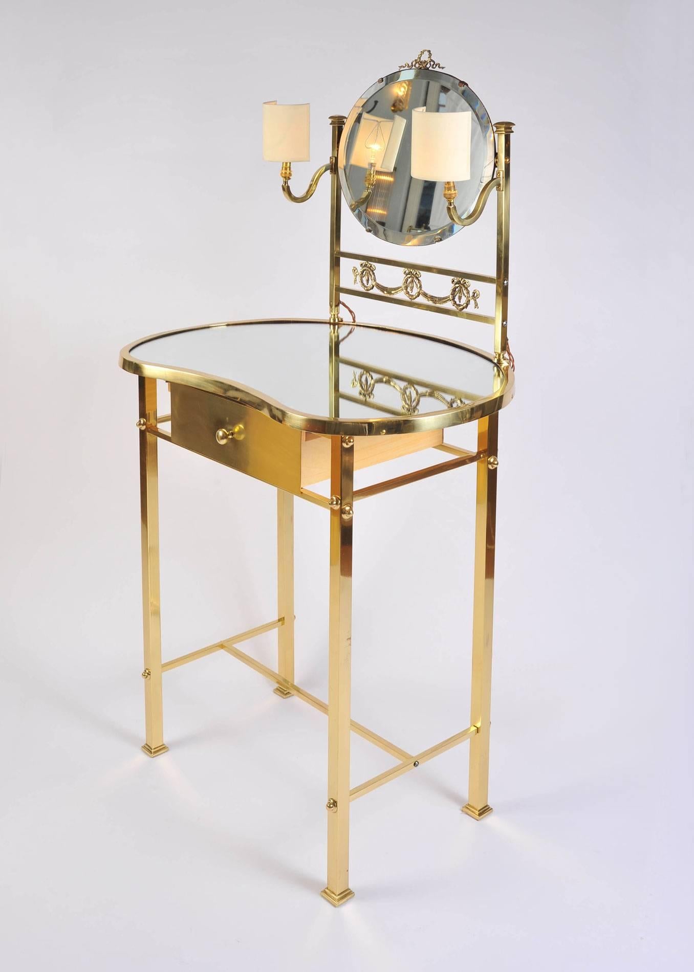 Kidney-shaped brass dressing table with brass drawer and mirrored top. Deep-beveled oval mirror supported by decorative brass frame with lamp holders.