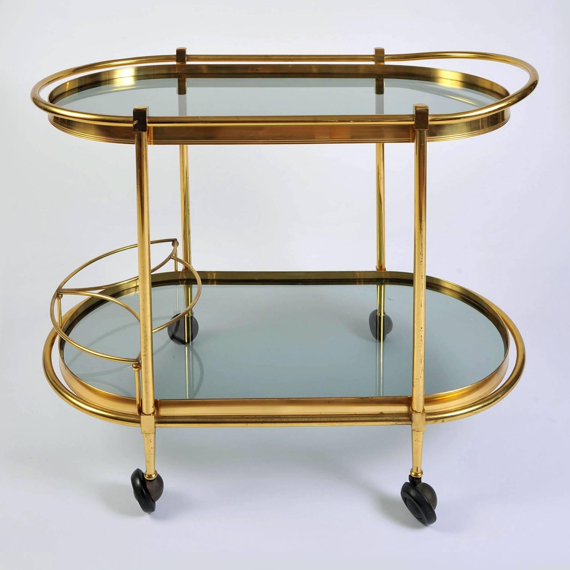 Curved bar cart/drinks trolley with two smokey glass shelves and bottle holder on wheels. Both shelves are surrounded by simple brass frames extending on the top shelf to create a handle.