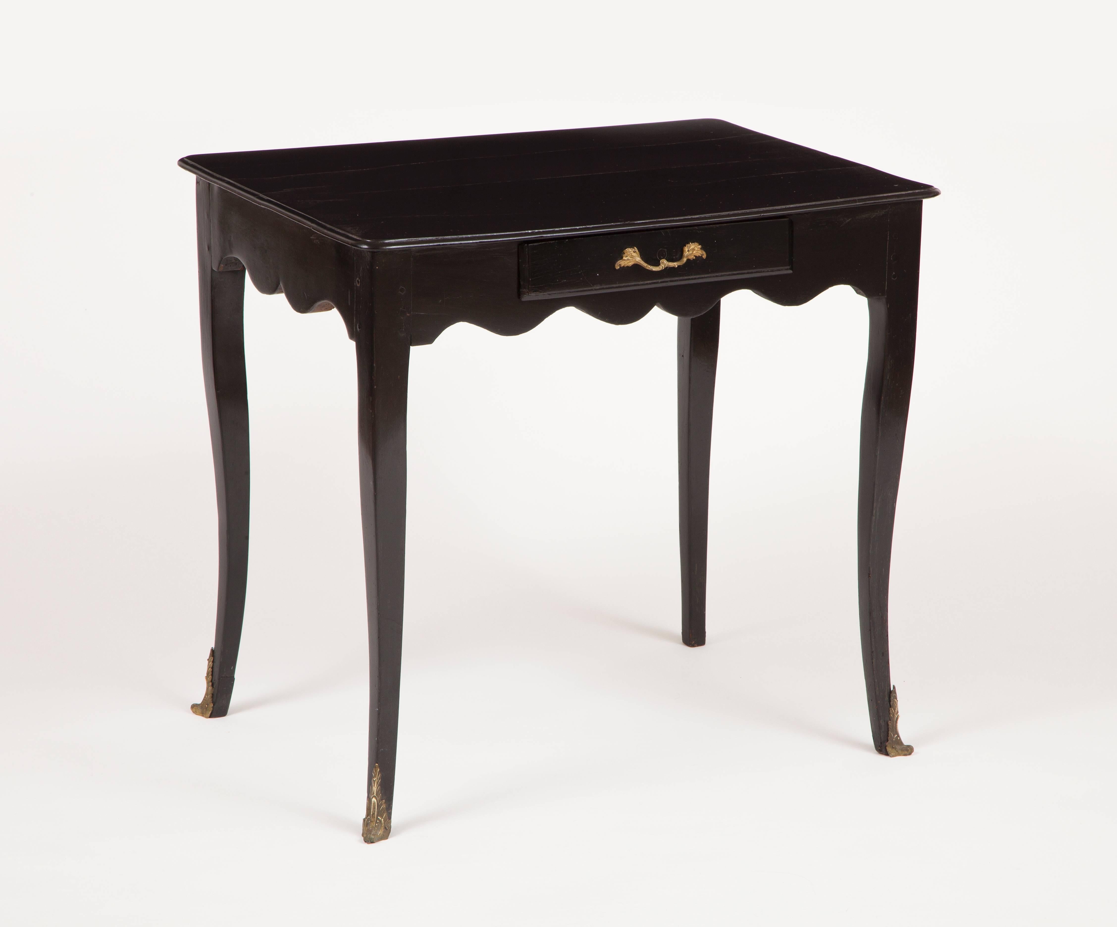 This French Louis XV ebonized wooden side table has a rectangular top with molded edge, above an undulating apron with one drawer fitted with gilded bronze pull, raised on curving cabriole legs, ending in gilded bronze sabots, the mounts of a later