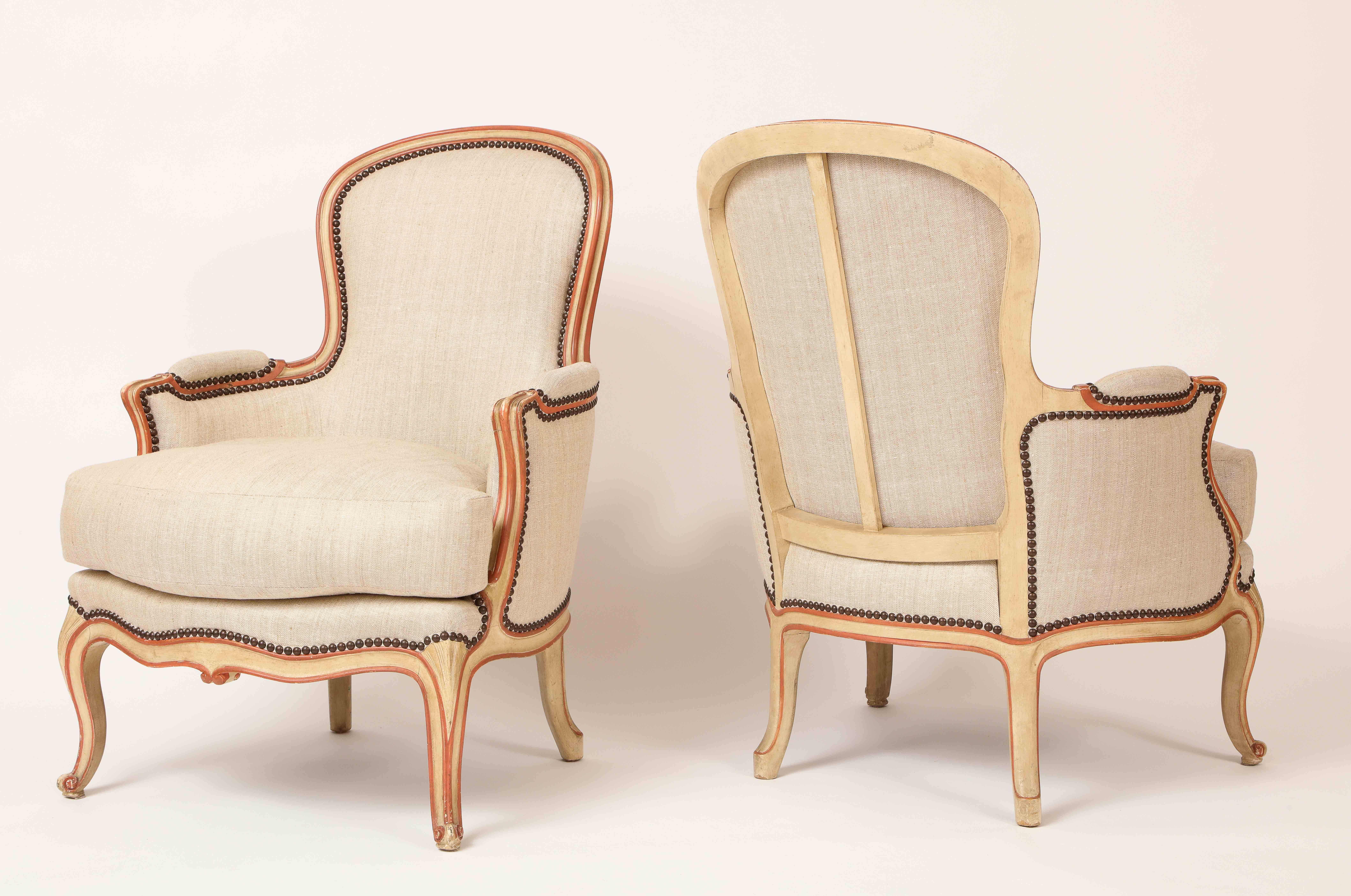 This pair of French armchairs (bergeres) in the Louis XV taste, made of painted beech wood, each has a curved back and arms, raised on cabriole legs, all retaining its original painted scheme of cream and orange pink. These chairs were made by