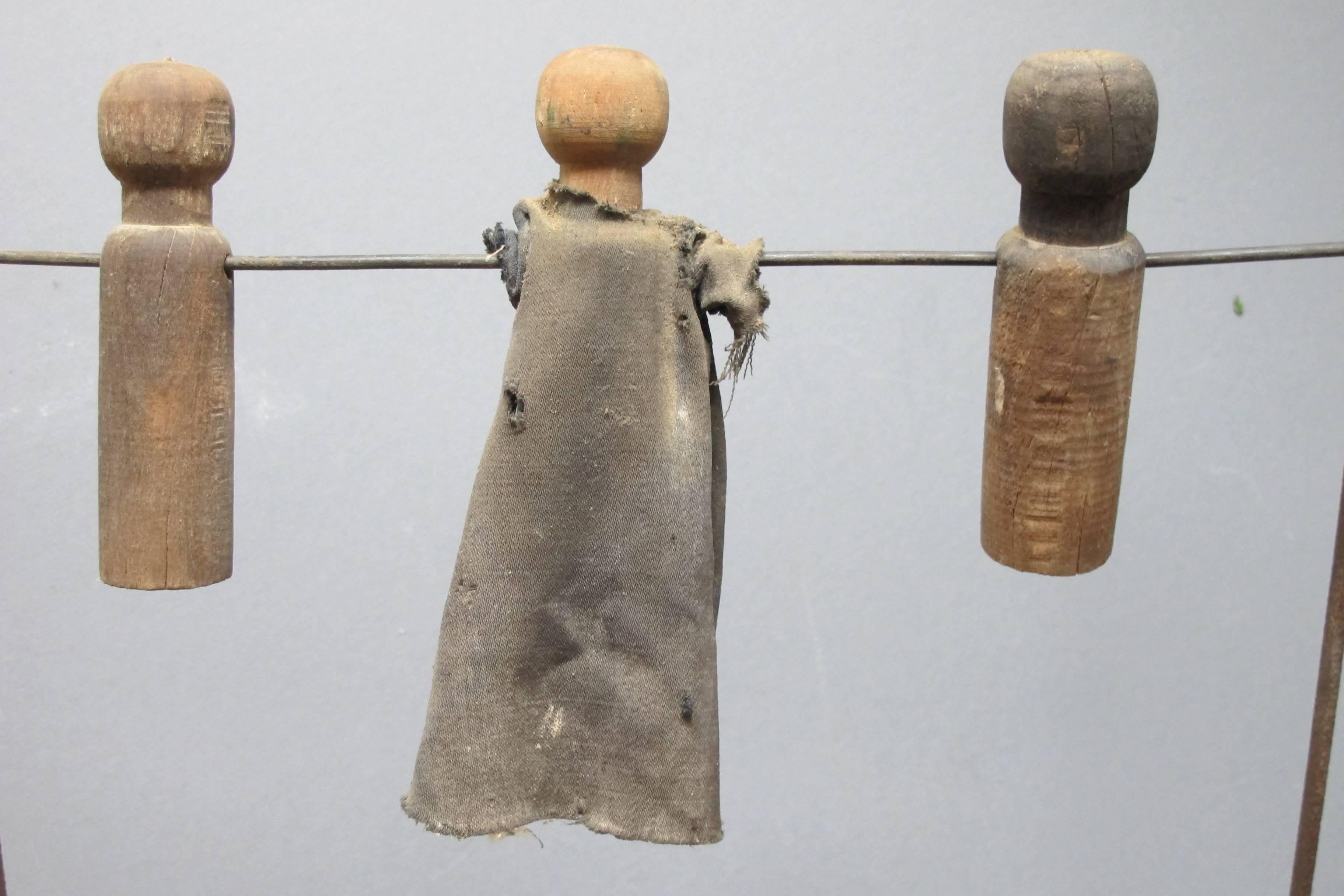 Metal frame with two horizontal rods hold three turned wood dolls with holes drilled through on each. Two of the figure have remains of their original clothes. I double frames pivots out to stand like a swing set or can be folded flat to hang on a