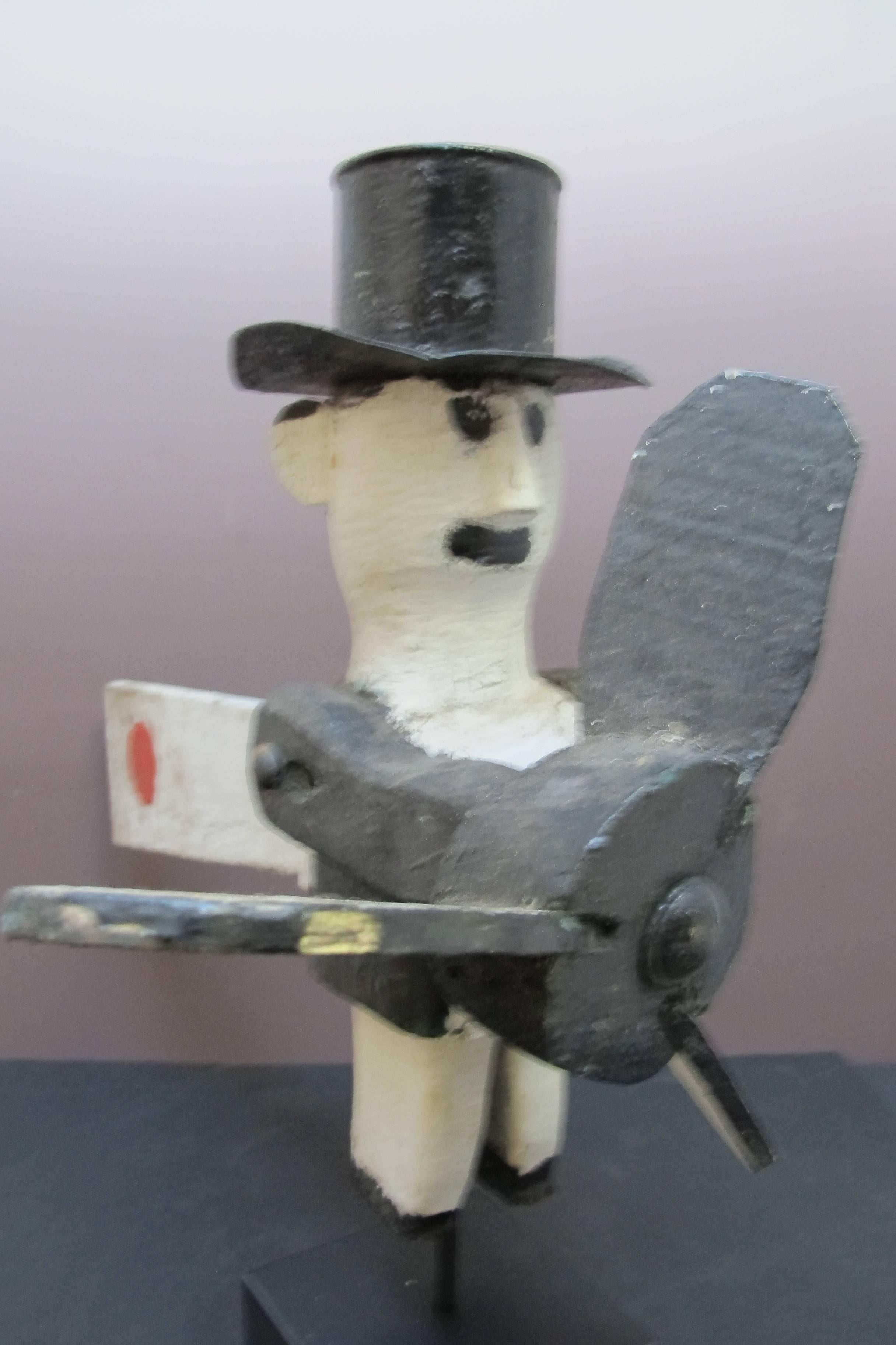 Unusual whirligig figure of a man with a tin top hat. The whirligig has one arm to the side and one extended forward holding a propeller. From the back a long wood tail extends with a painted red dot. The figure shows earlier paint with a great