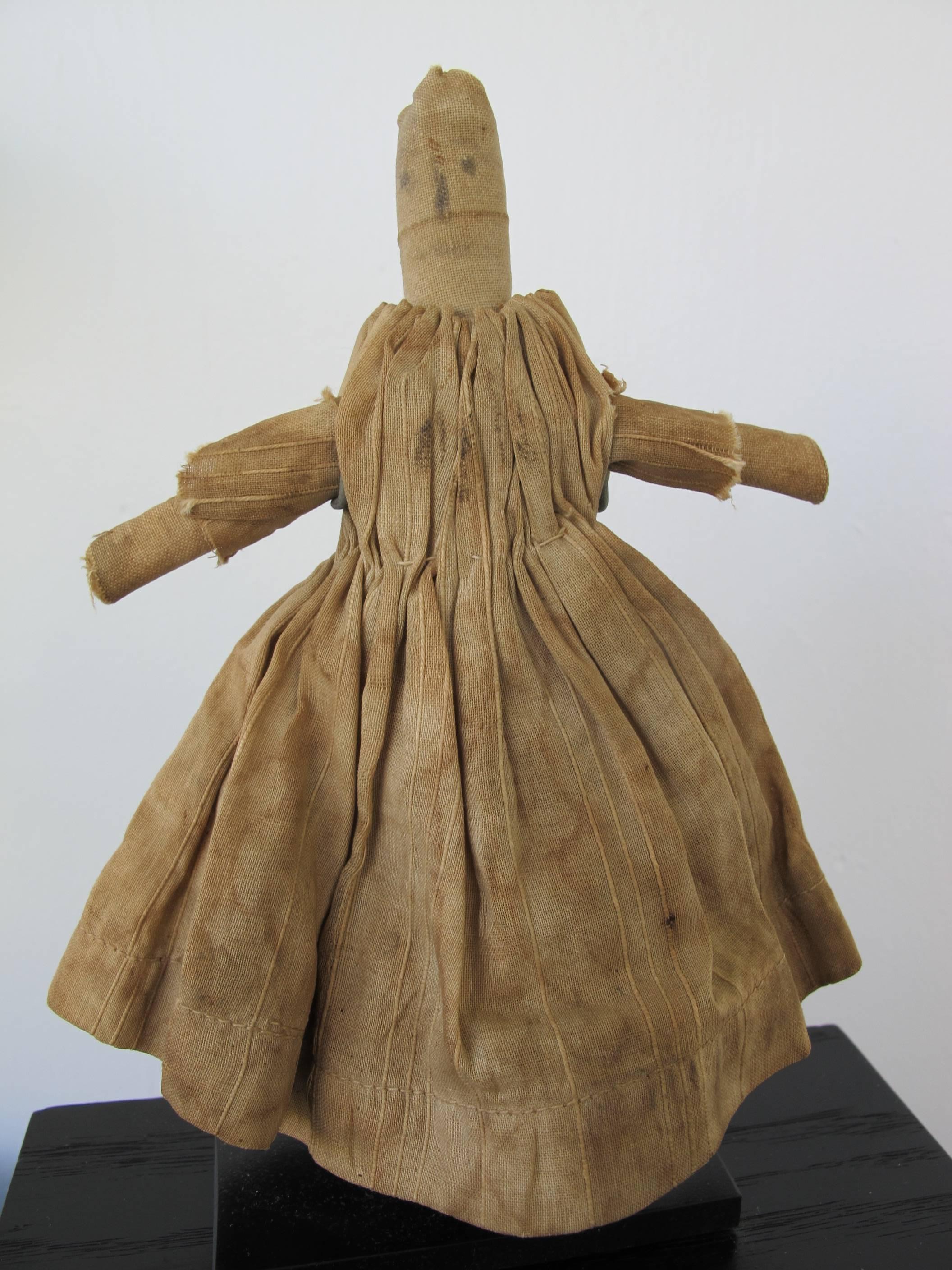 Simple cloth doll of homespun linen with rolled cloth raised arms, legs and head.
The gathered dress was once blue and is now a mottled tan. Mounted on metal base. Pictured in American Folk Dolls by Wendy Lavitt. illus. page 21, full page.
