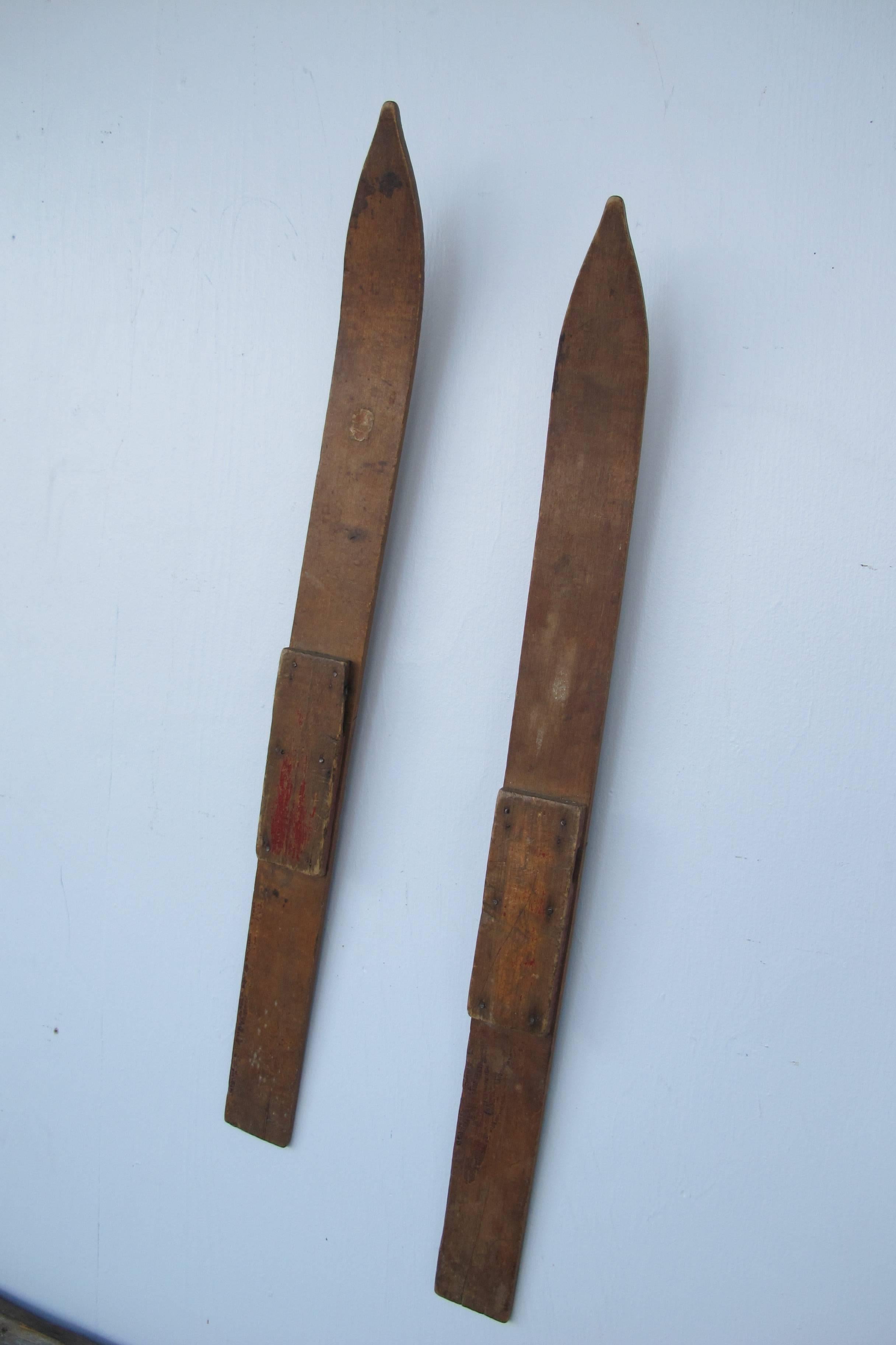 Diminutive wooden skis with remains of some reddish wash. They look great at an angle on the wall. The bentwood skis taper to a raised point and have a center groove on the bottoms. A rectangular strip of wood is nailed on where the boot would be