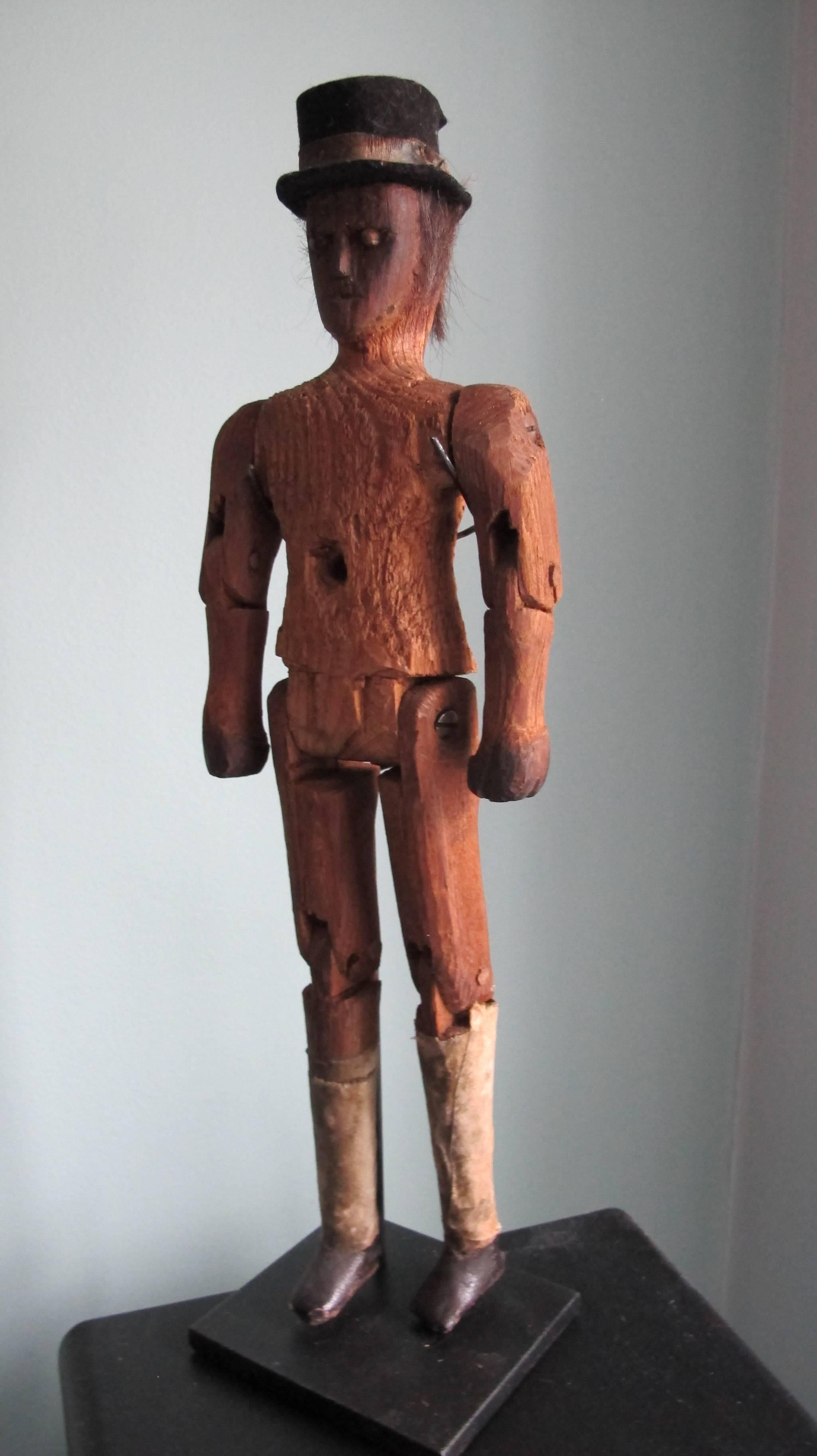 Carved wood figure with moveable arms and legs and hole in chest for inserting a stick to make it walk or dance. The head has bead eyes with pin centres and cloth top hat and hair. The black painted shoes have knickers above of adhered cloth. There