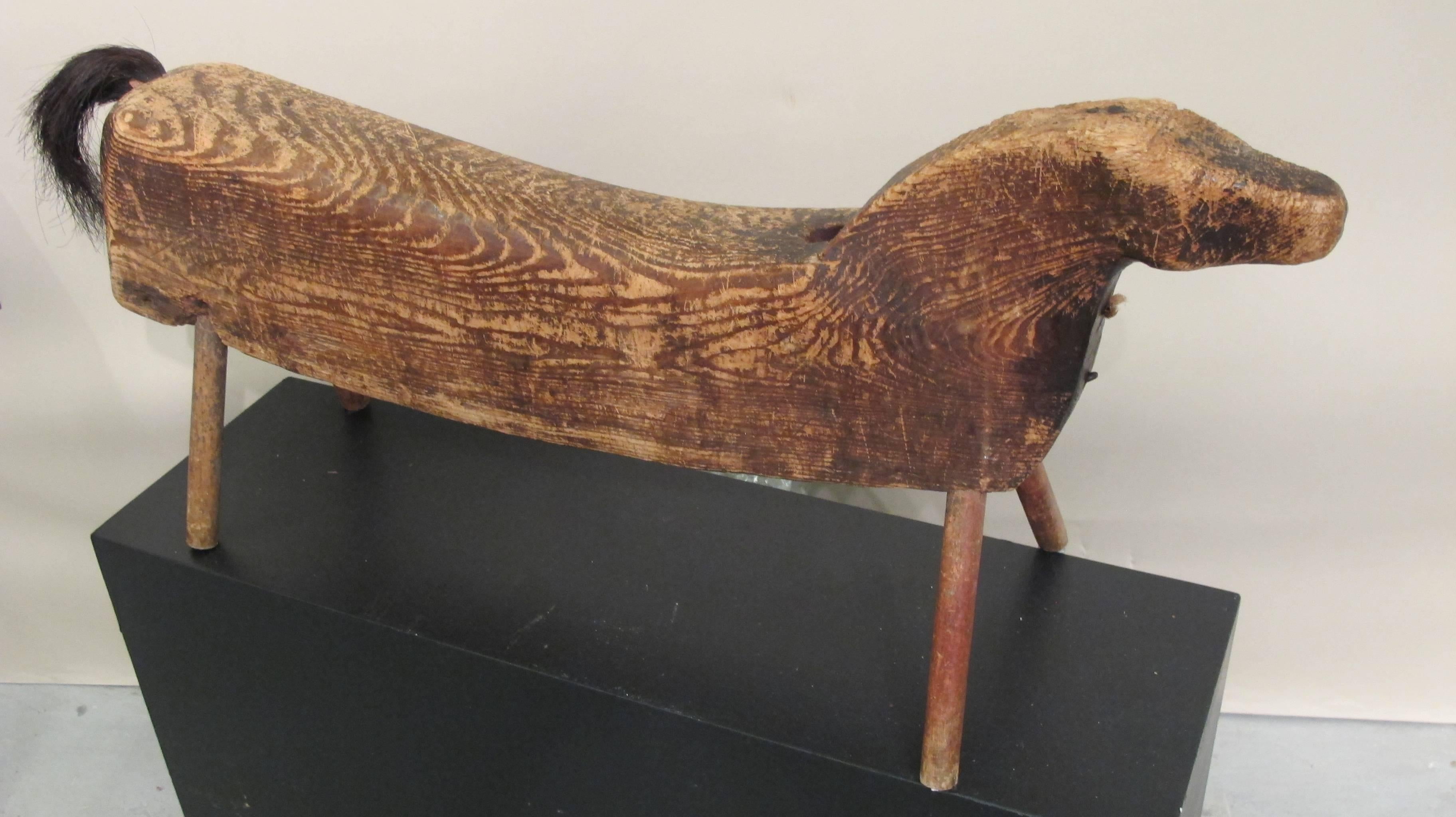 Carved wood horse with elongated body and horse tail. The young child would sit and ride on the horse supported by angled wood peg legs. The horse is a piece of sculpture by an unknown folk artist of the 19th century.
