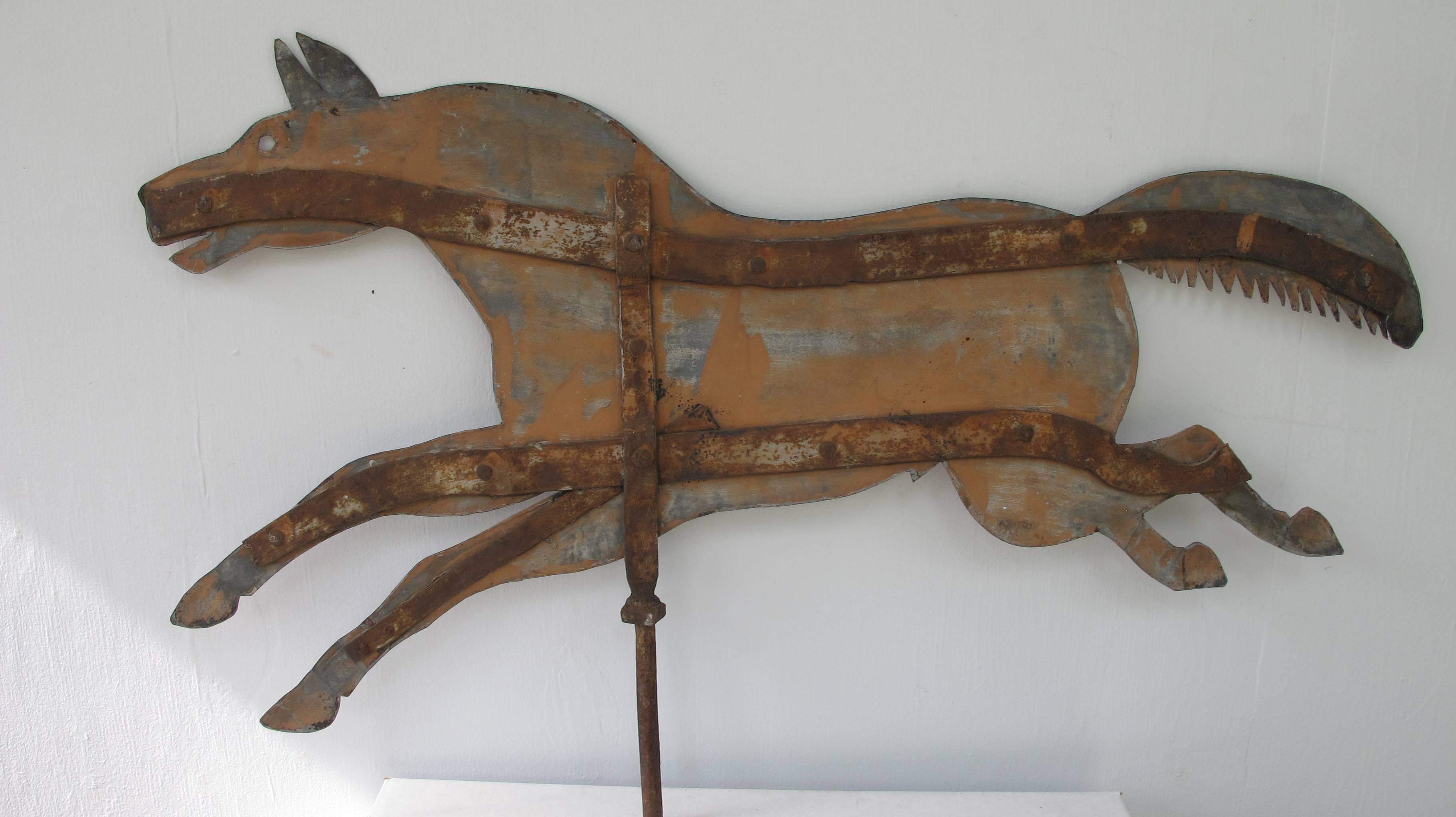 Handmade running horse weathervane of cut sheet metal with iron bracing on one side.
The weathered surface has remains of old paint on the zinc surface. The original iron rod is attached that would have attached it to the roof to turn with the