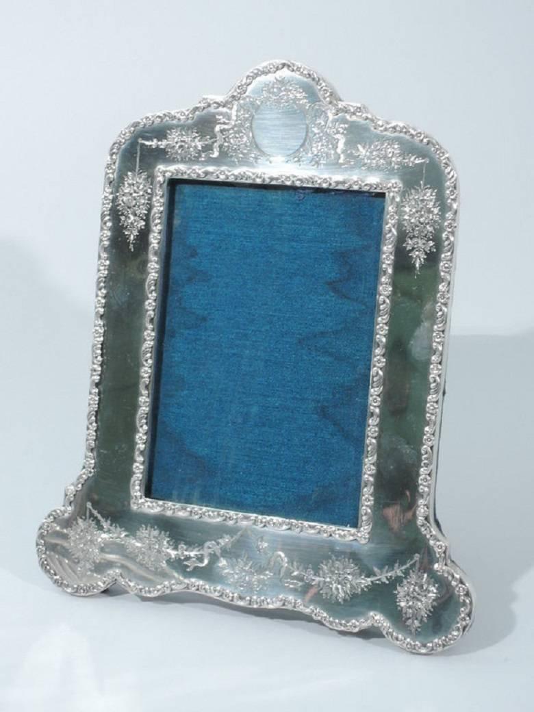 Edwardian sterling silver picture frame. Made by William Adams in Birmingham in 1904. Straight sides, stepped and arched top, and scalloped bottom. Top corners are round. Bottom corners lobed. Applied scrolls and flowers border window and frame.