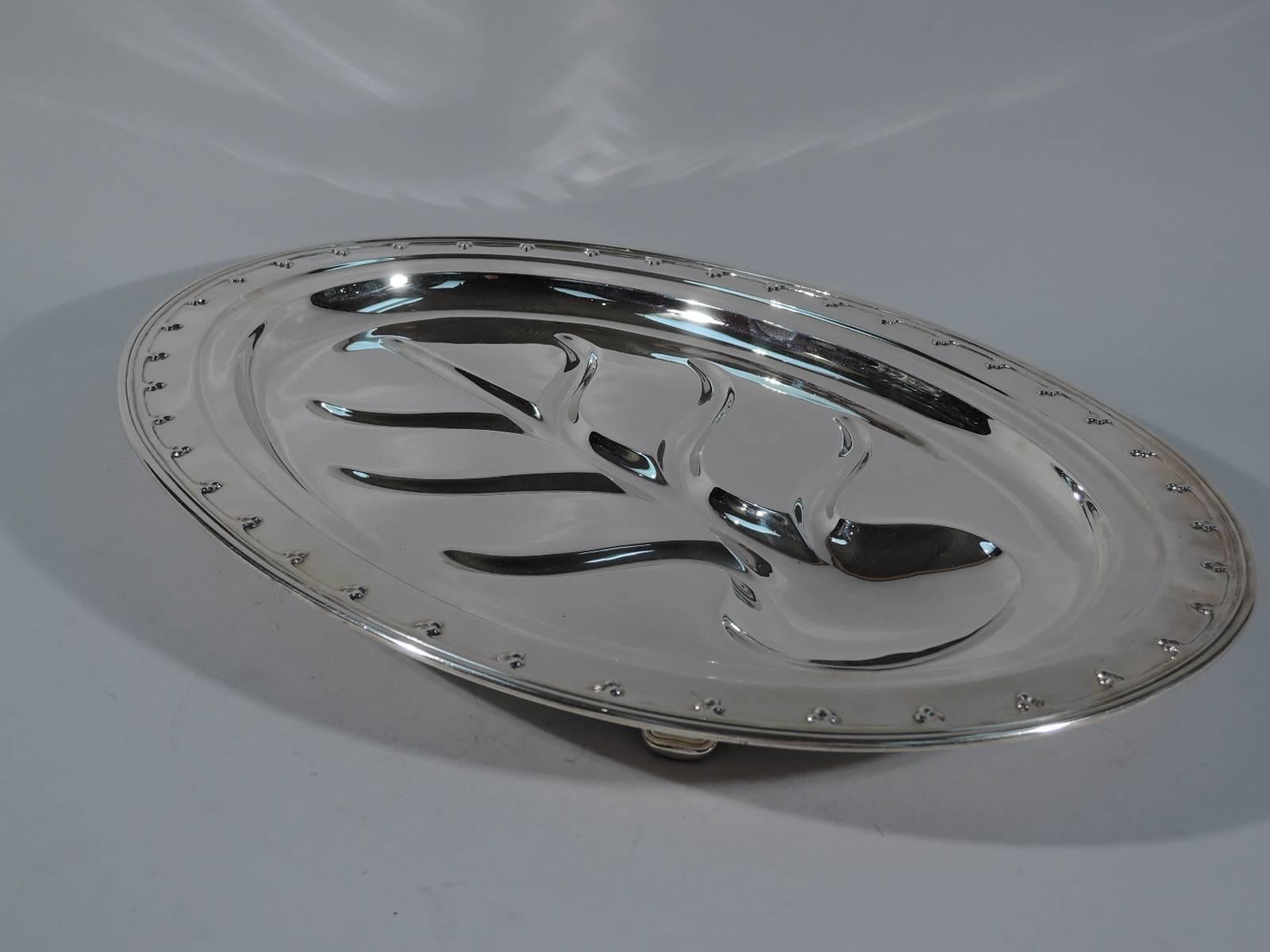 Sterling silver meat platter in Saint Dunstan pattern. Made by Tiffany & Co. in New York, ca 1921. Oval with applied fleur-de-lys rim. Well has traditional Tree of Life “canals” to collect the rivulets of precious pan juices. Rests on 4 supports. A