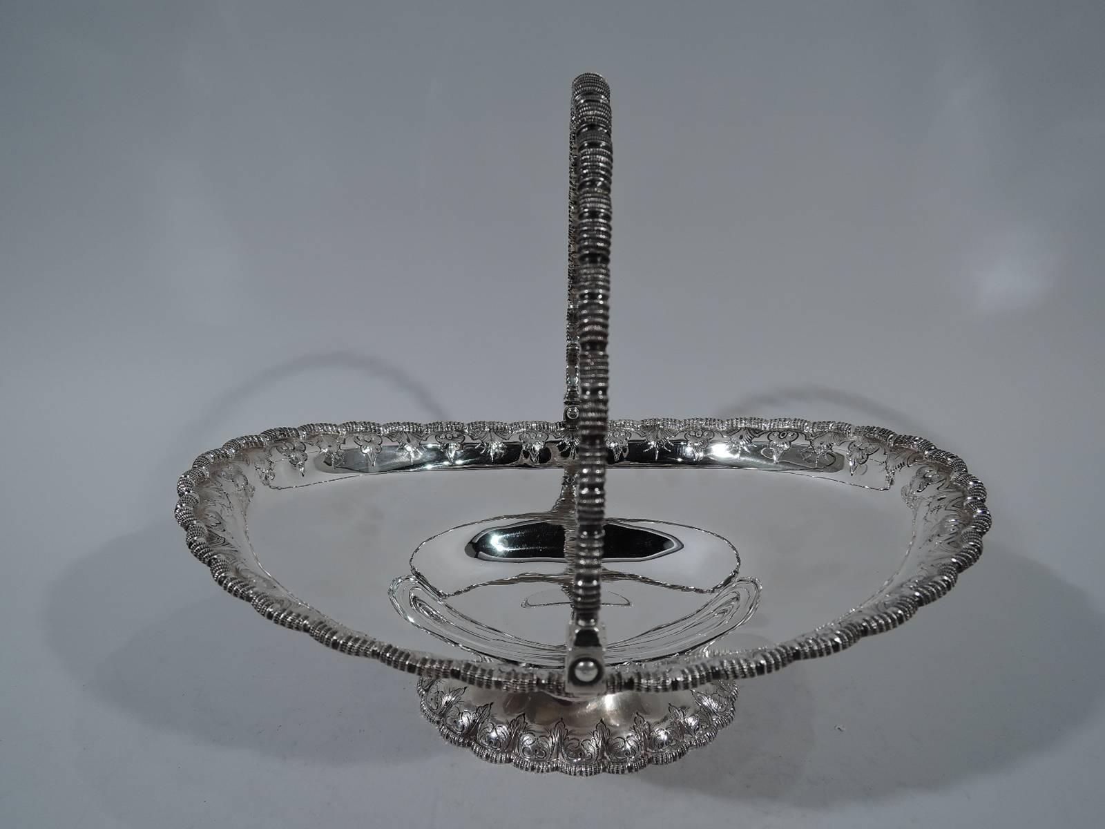 Early sterling silver basket. Retailed by Tiffany & Co. at 550 Broadway, New York. Oval shallow bowl and scalloped gadrooned rim with chased and engraved fleurs de lys. Foot raised and lobed with scalloped gadrooned rim and engraved leaves. Swing