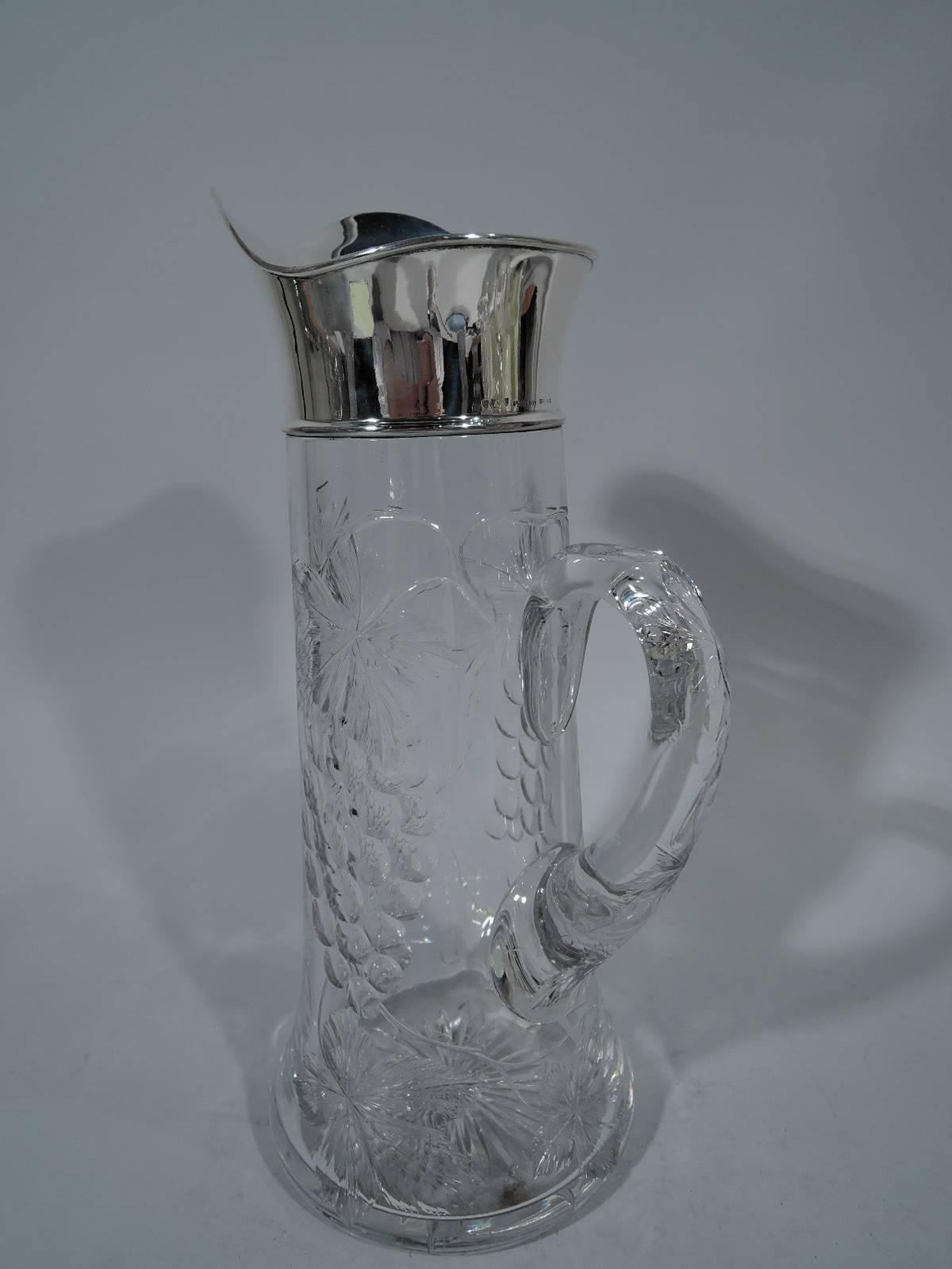 Glass claret jug with sterling silver collar. Made by Shreve & Co. in San Francisco, circa 1910. Cylindrical with spread base and scroll handle. Fruiting grapevine with big leaves and succulent fruit bunches cut to body and handle. Glass clear to