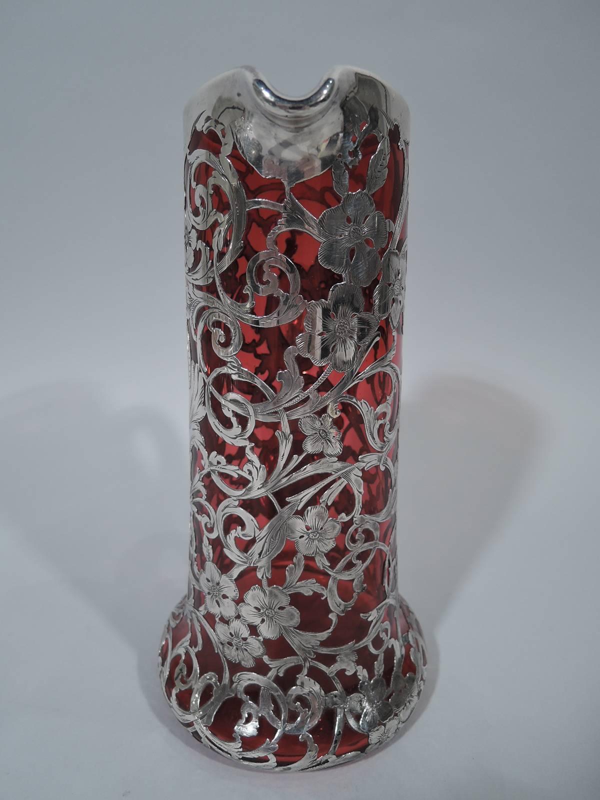 Art Nouveau red glass claret jug with silver overlay, circa 1900. Cylindrical with small lip spout, spread base, and scroll handle. Dense silver scrolls and flowers as well asymmetrical cartouche with engraved monogram. Mouth and handle have silver