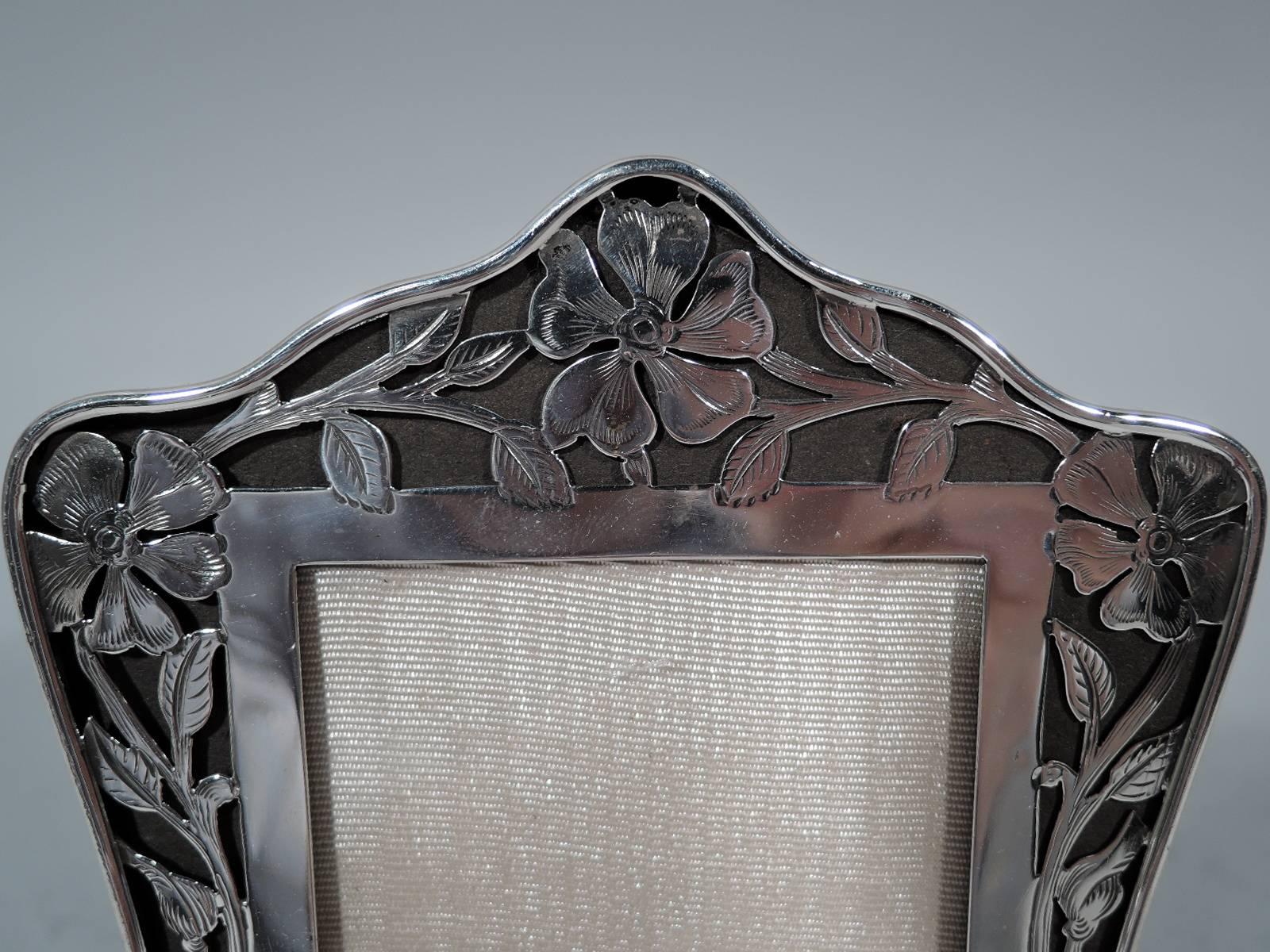 Art Nouveau sterling silver picture frame. Rectangular window in shaped frame with wavy arched top, concave sides, and bracket feet. Window surrounded by pierced floral border over brown ground. At bottom vacant oval. With glass, silk lining, and