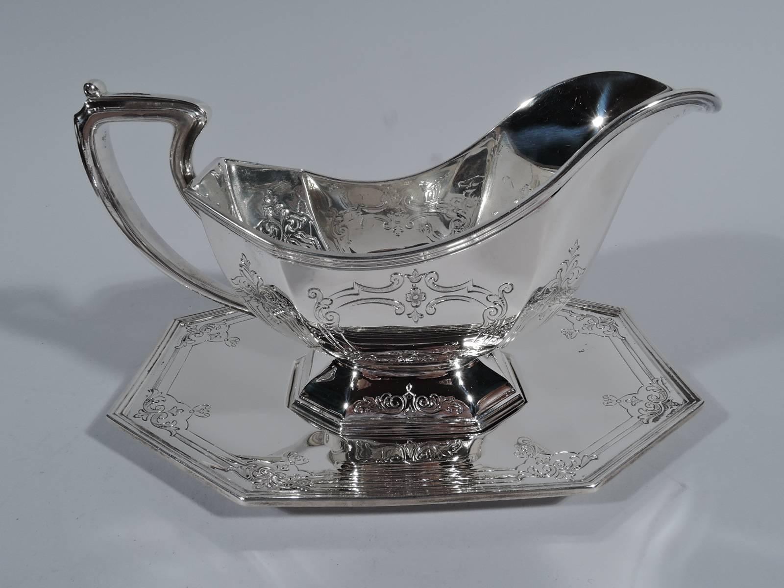 Sterling silver gravy boat on stand. Made by Reed & Barton in Taunton, Mass., circa 1910. Chamfered corners and reeded rim. Boat has helmet mouth, domed foot, and capped scrolled bracket handle. Scrolled and lined frames with stylized flowers and