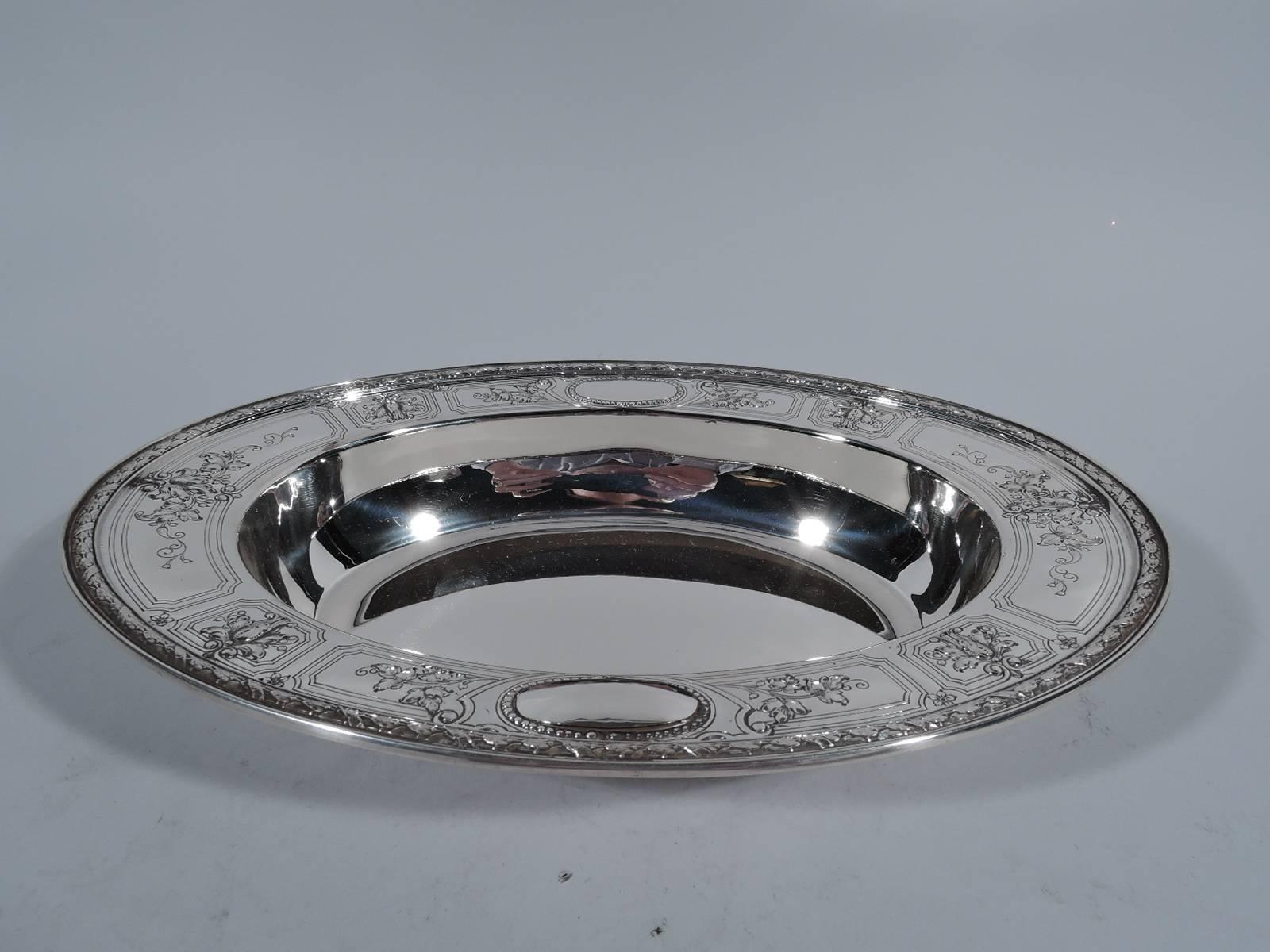 Fancy sterling silver serving bowl. Made by Gorham in Providence in 1928. Plain oval bowl with wide and flat rim. Rim has shaped frames inset with raised flowers that spill over the lines. Two raised ovals with beaded rims (vacant). Imbricated