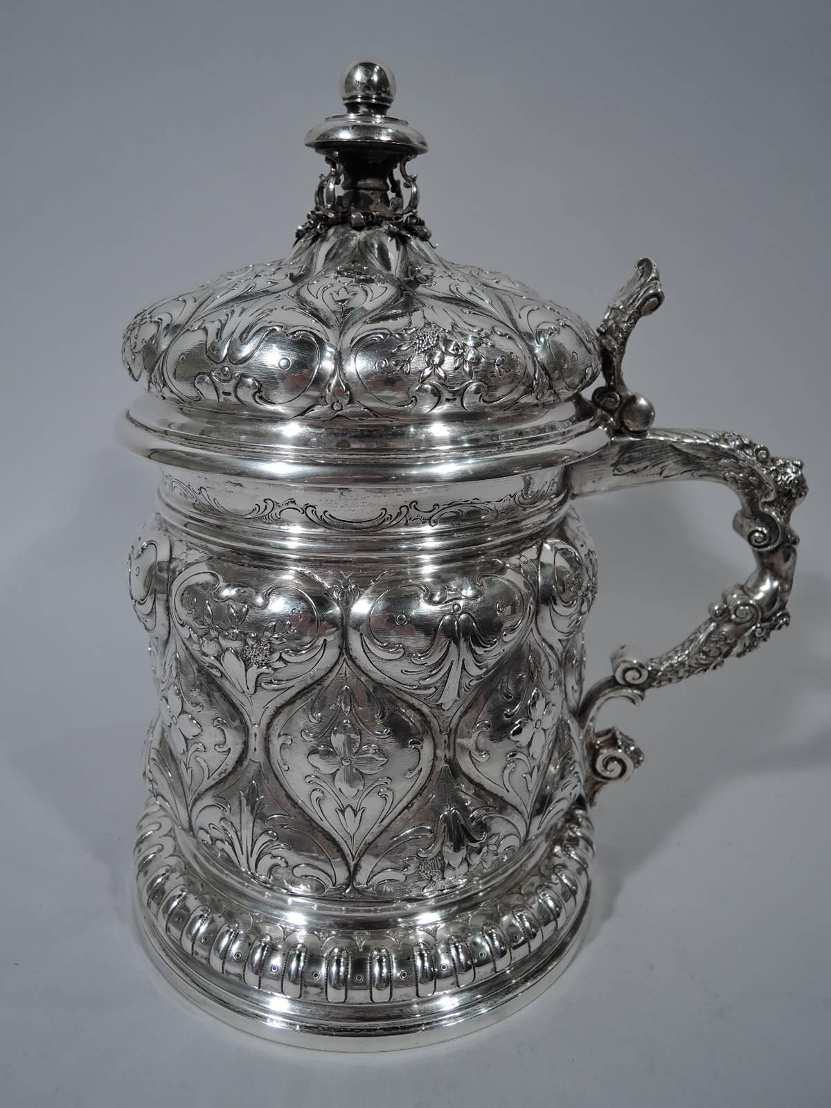 Antique German 800 silver tankard in 18th century style. Drum-form bowl with raised foot, hinged and domed cover, and s-scroll finial. Floral-decorated quilting, hinged bun cover with mask thumb rest and ornamental finial, and double-scroll handle