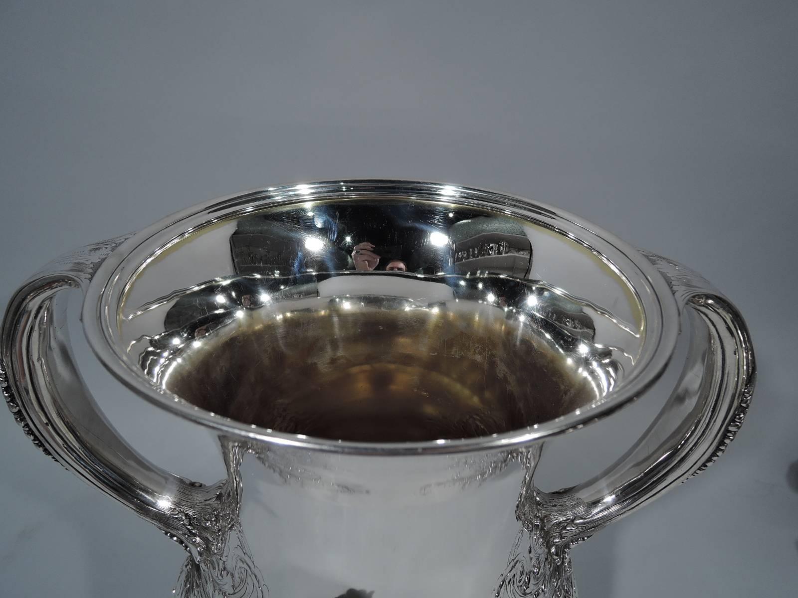 Large and heavy sterling silver trophy cup. Made by Tiffany & Co. in New York. Baluster bowl with raised foot, c-scroll handles, flared rim, and slashed lobbing. Handles reeded and leaf-capped, and have feathery leaf mounts. Gilt-washed interior. A