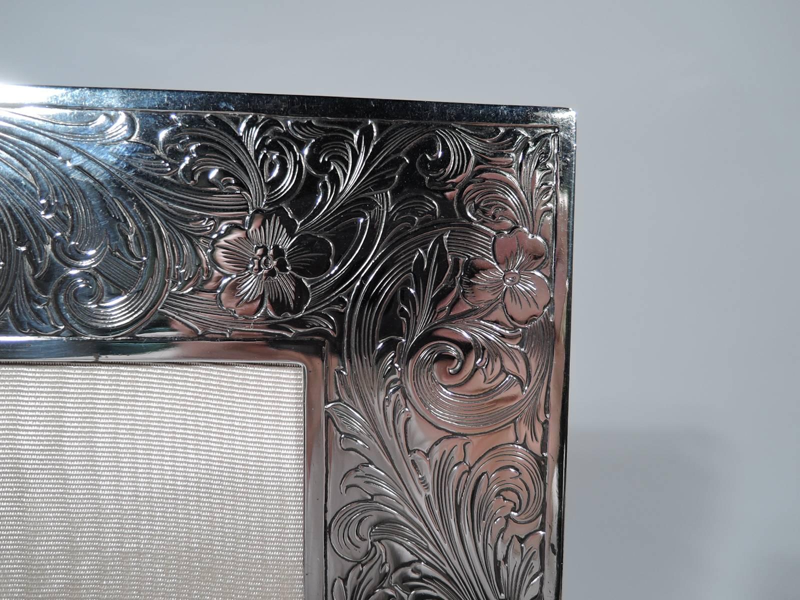 Edwardian sterling silver frame. Made by Gorham in Providence in 1914. Rectangular window and wide flat border engraved with dense and dynamic flowers and scrolls. Scrolled cartouche (vacant). With glass, silk lining, and velvet back and hinged