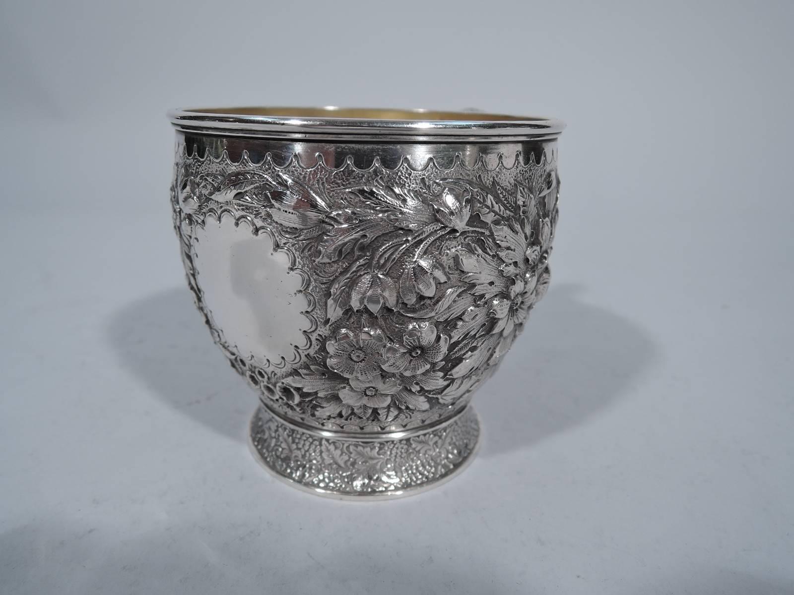 Sterling silver baby cup. Made by Tiffany & Co. in New York, circa 1875. Curved sides, spread foot, and scroll foot. Body has dense and naturalistic floral repoussé on stippled ground, scalloped circular frame (vacant), and more scalloping at top