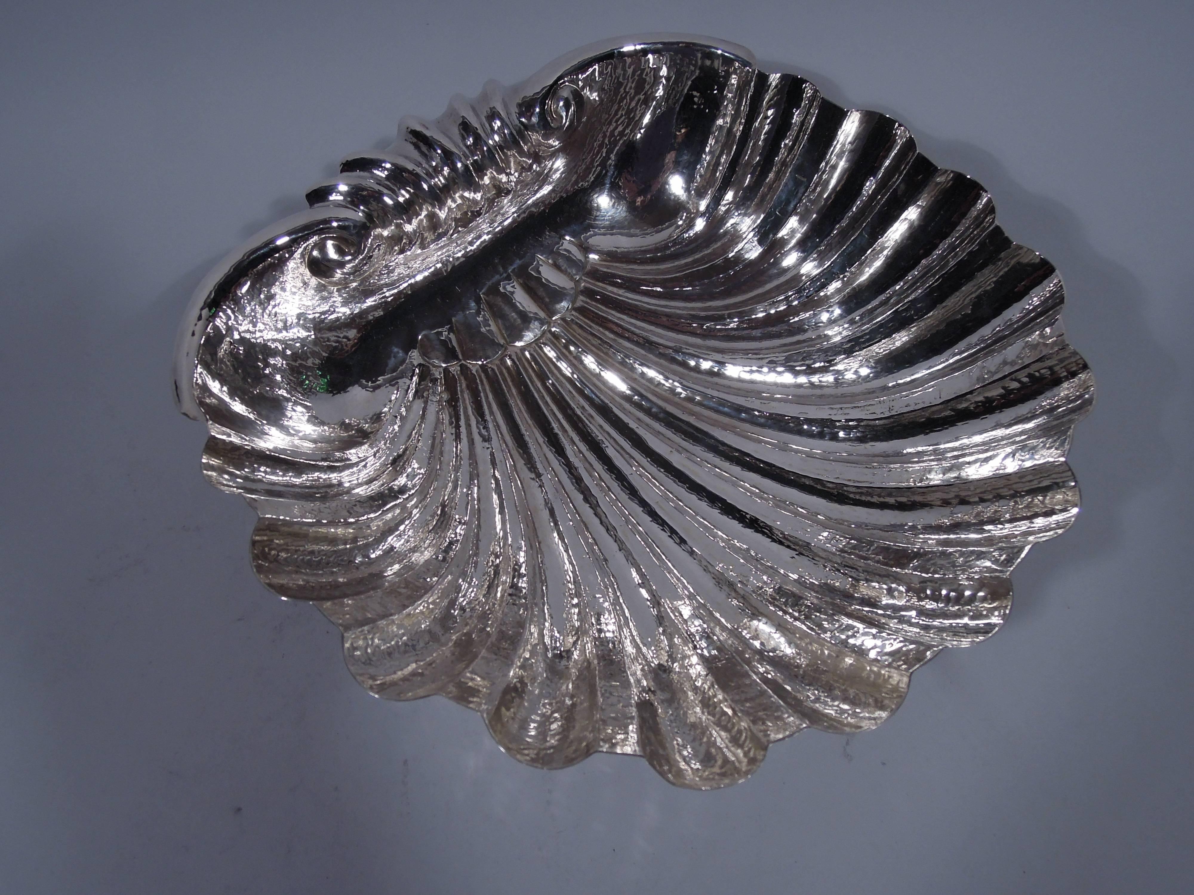 Sterling silver scallop shell bowl. Deep with flutes and curvilinear scalloped rim. Scrolled handle bordered by volute scrolls. Underside has incised hatching and three seashell supports. Unusual and textural with visible hand hammering. Hallmarked