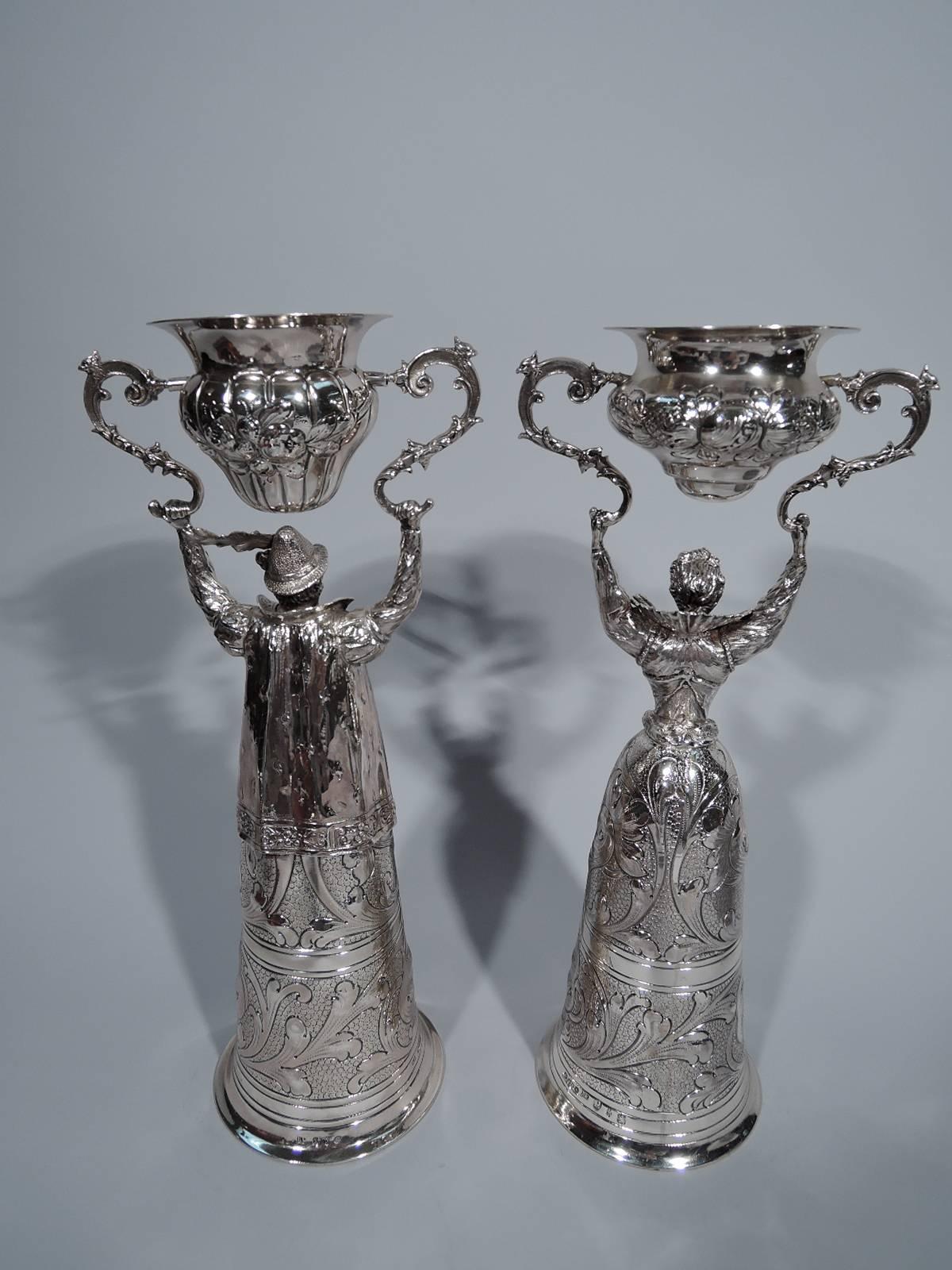 A wonderful pair of German sterling silver marriage (or wager) cups. This pair comprises a man and woman in Renaissance attire, each holding overhead two scrolled brackets with swing cup. Fantastic details including lace ruff and embroidered bodice