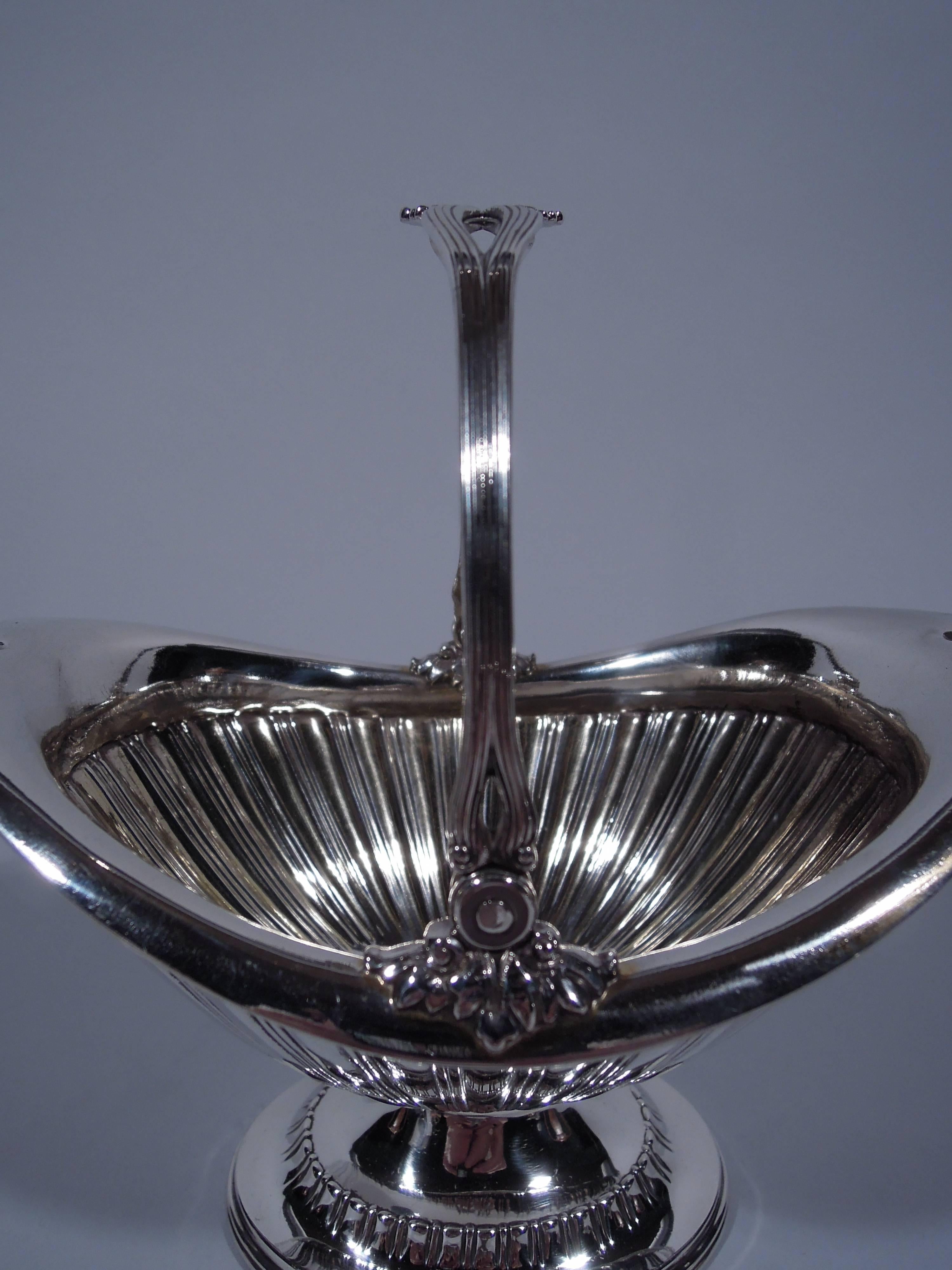 Sterling silver sugar basket. Made by Tiffany & Co. in New York, 1892-1902. Ovoid with asymmetrical rim, short stem, and stepped oval foot. Sides have alternating lobes and double lines. Reeded swing handle with leaf mounts. Hallmark includes