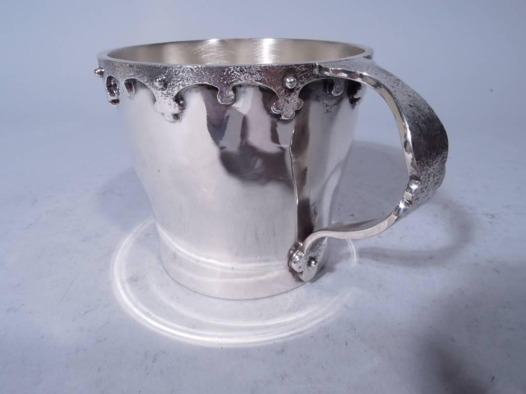 Arts & Crafts sterling silver mug. Made by JE Caldwell in Philadelphia, circa 1915. Gently tapering body. Hand-hammered and studded strapwork mounted to rim. Scroll handle also hand-hammered with studded trefoil terminal. Unusual for a baby cup it’s